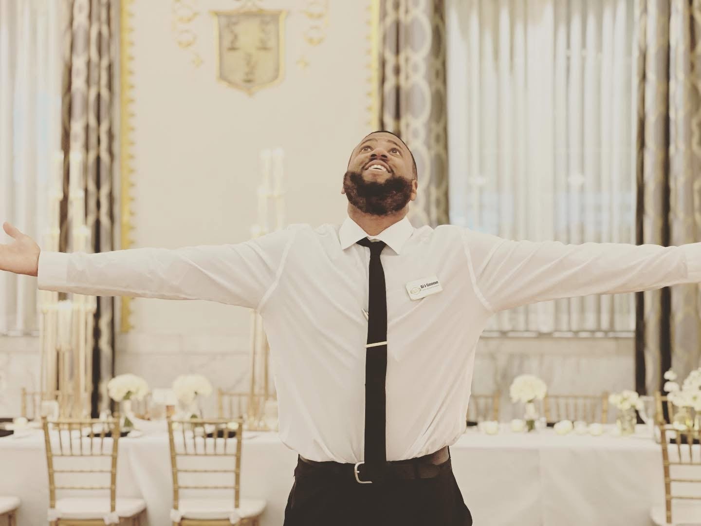 Man with beard in formal attire with arms open, in a decorated room with white and gold table settings and floral arrangements.