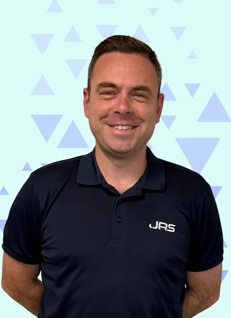 A smiling man with short brown hair, wearing a black polo shirt with 'JRS' embroidered on it, standing in front of a light blue background with a pattern of darker blue triangles.