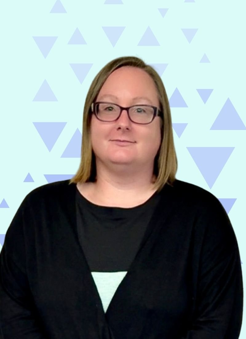 A woman with shoulder-length blonde hair, wearing glasses and a black top, standing against a light blue background with a pattern of light blue triangles.