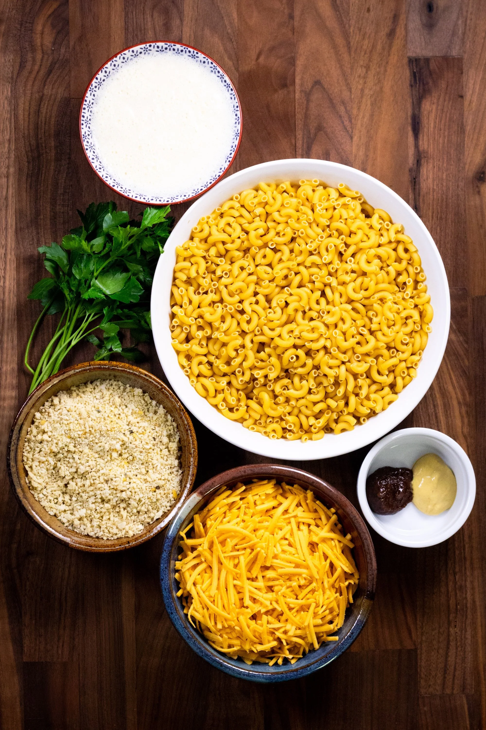 Bowls of elbow pasta, vegan cheddar, breadcrumbs, and sauce ingredients on a wooden board.