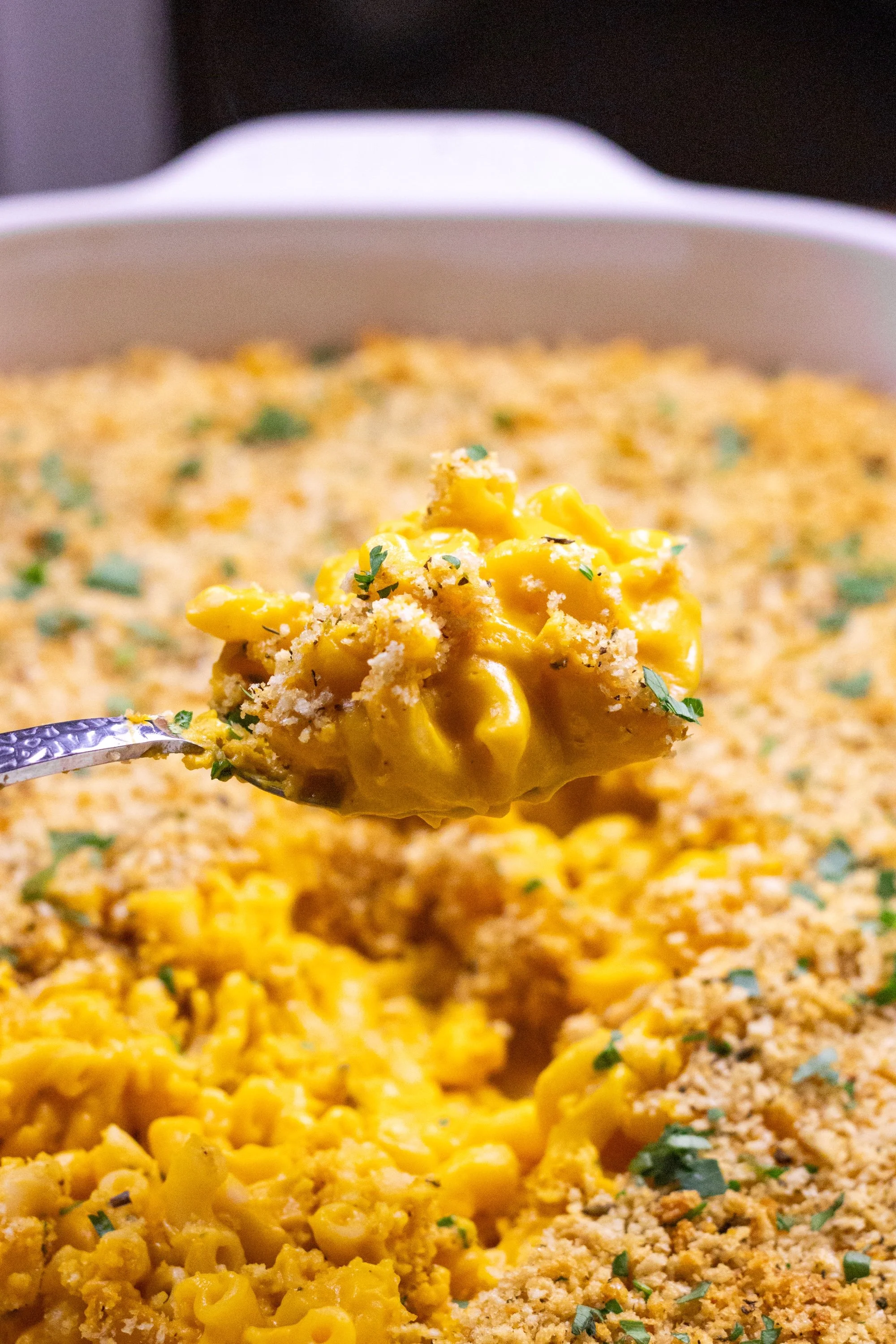A spoonful of creamy vegan butternut squash mac and cheese with toasted breadcrumbs and parsley, held above the baking dish.