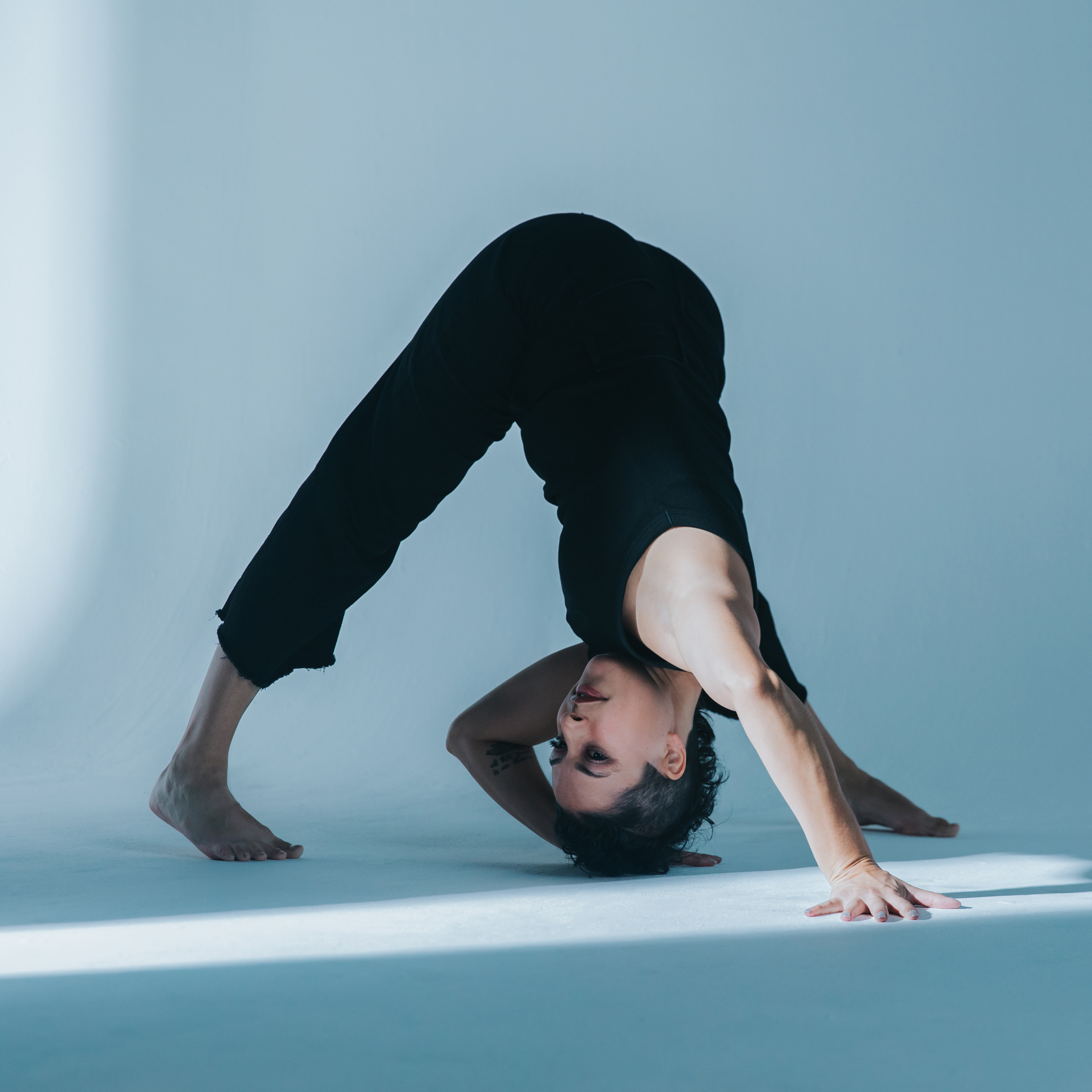 Person performing a yoga pose on the floor, head upside down, knees bent, wearing black clothes, with a neutral background.