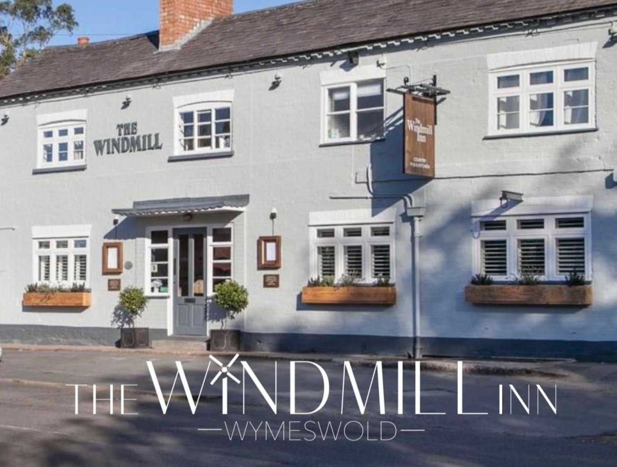 Front exterior of The Windmill Inn in Wymeswold, a white two-story building with multiple windows, potted plants, and a sign hanging outside.