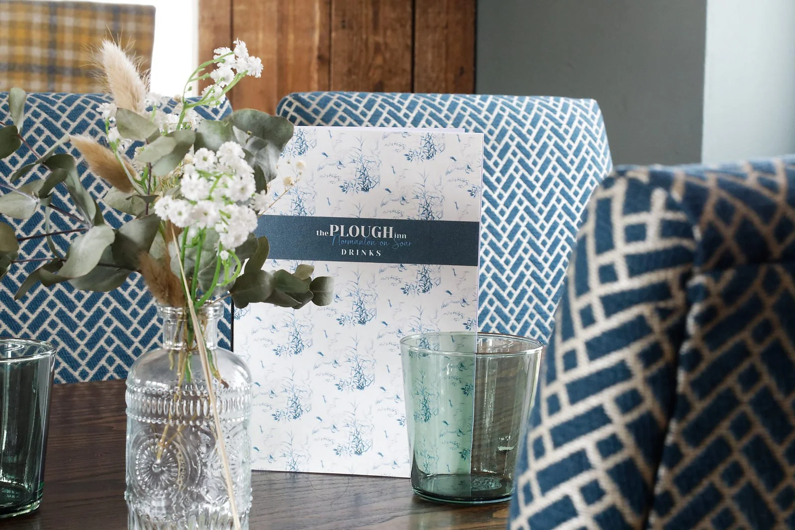 A floral arrangement in a clear textured glass vase placed on a wooden table, with a menu or brochure behind it reading 'the PLOUGH Inn' and 'DRINKS'. There are also empty glasses on the table, and patterned blue and cream upholstered chairs surrounding the table.