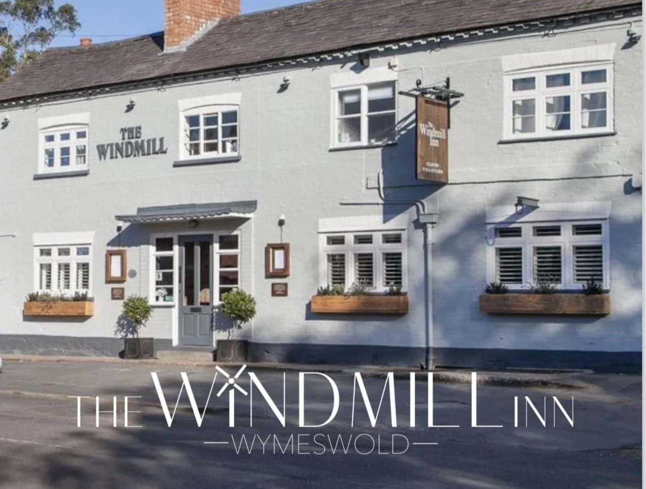 Exterior view of The Windmill Inn, a guesthouse with a white facade, multiple windows with shutters, wooden flower boxes, and a sign hanging outside.