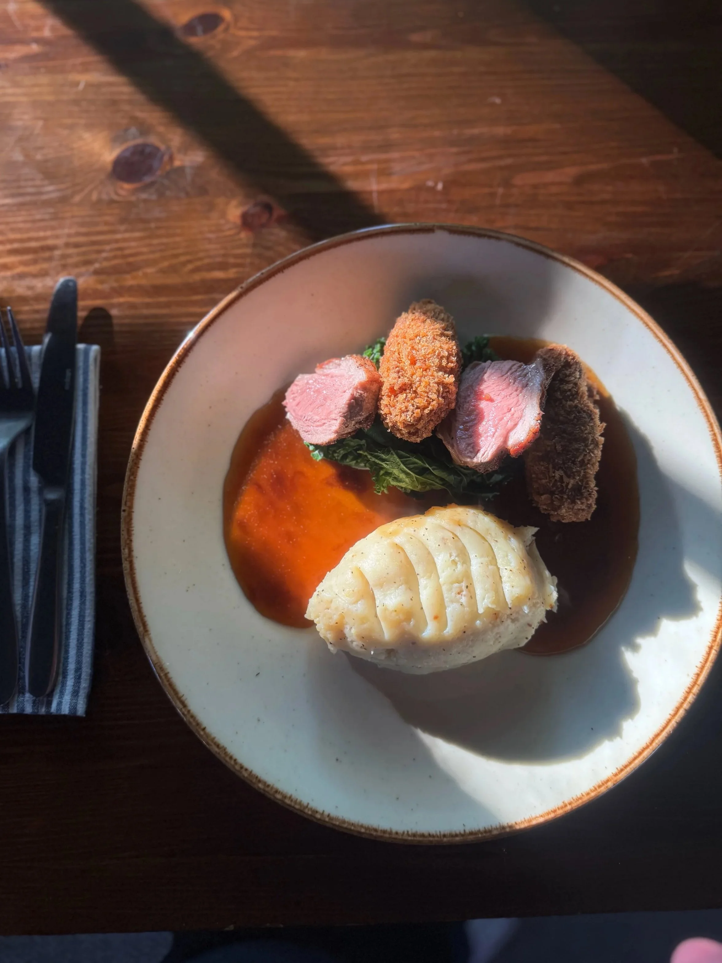 A plate of food with three pieces of fried or cooked meat on a bed of greens, a mound of creamy mashed potatoes with grill marks, and a pool of brown gravy on a white ceramic plate placed on a wooden table.