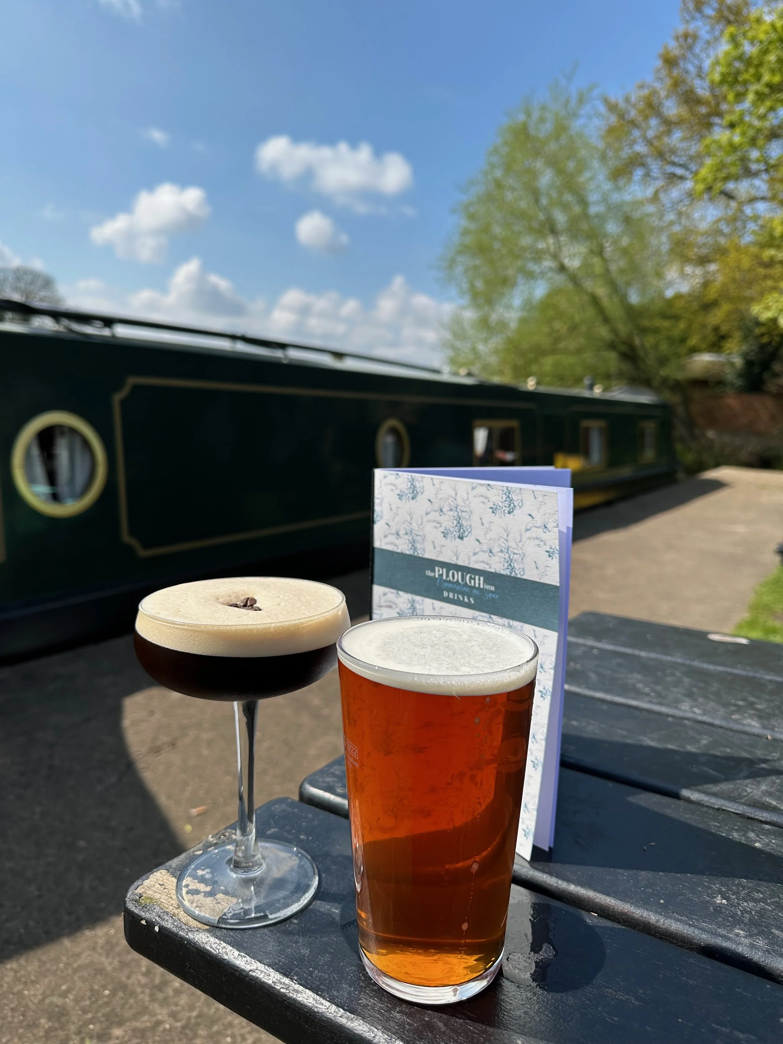 A black and tan beer in a glass with a foamy head, and a dark cocktail with a foamy top and coffee bean garnish, on a black wooden outdoor table. A menu or brochure and a canal boat are in the background, with trees and a partly cloudy sky.