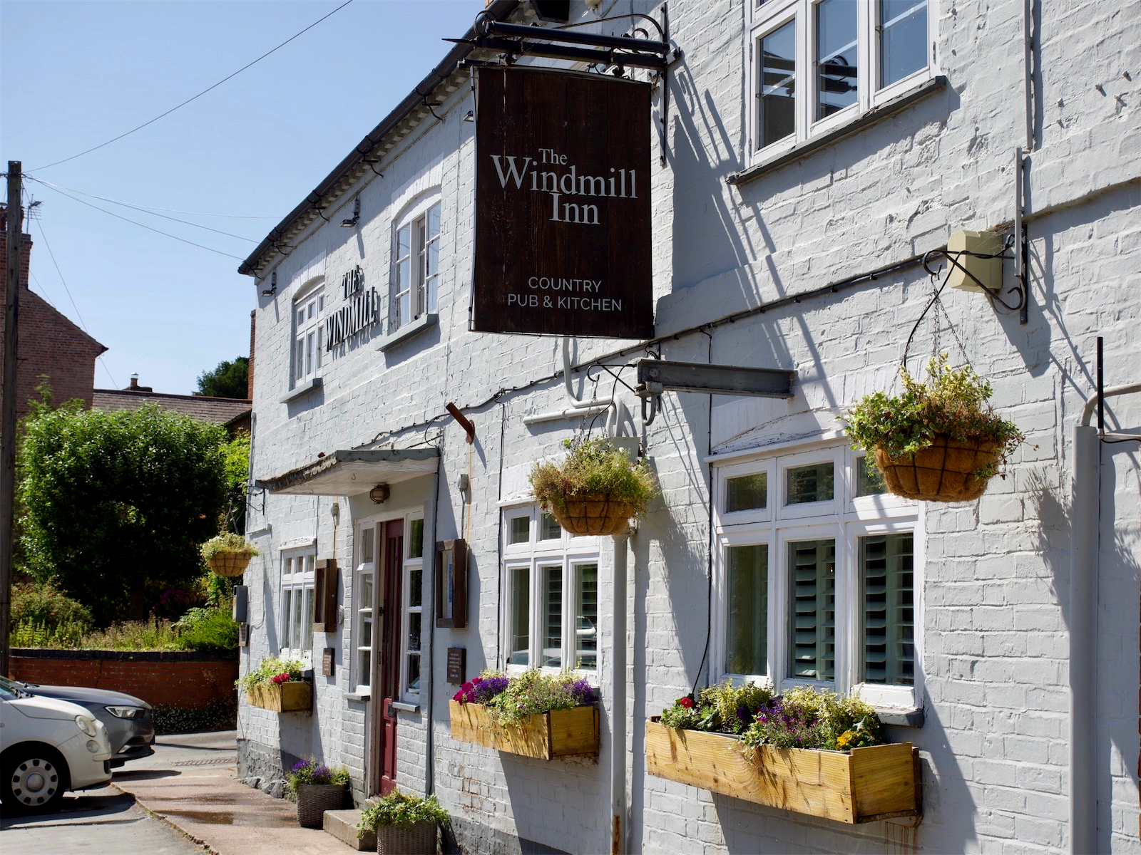 White brick building with hanging flower baskets and flower boxes, sign reading "The Windmill Inn, Country Pub & Kitchen."