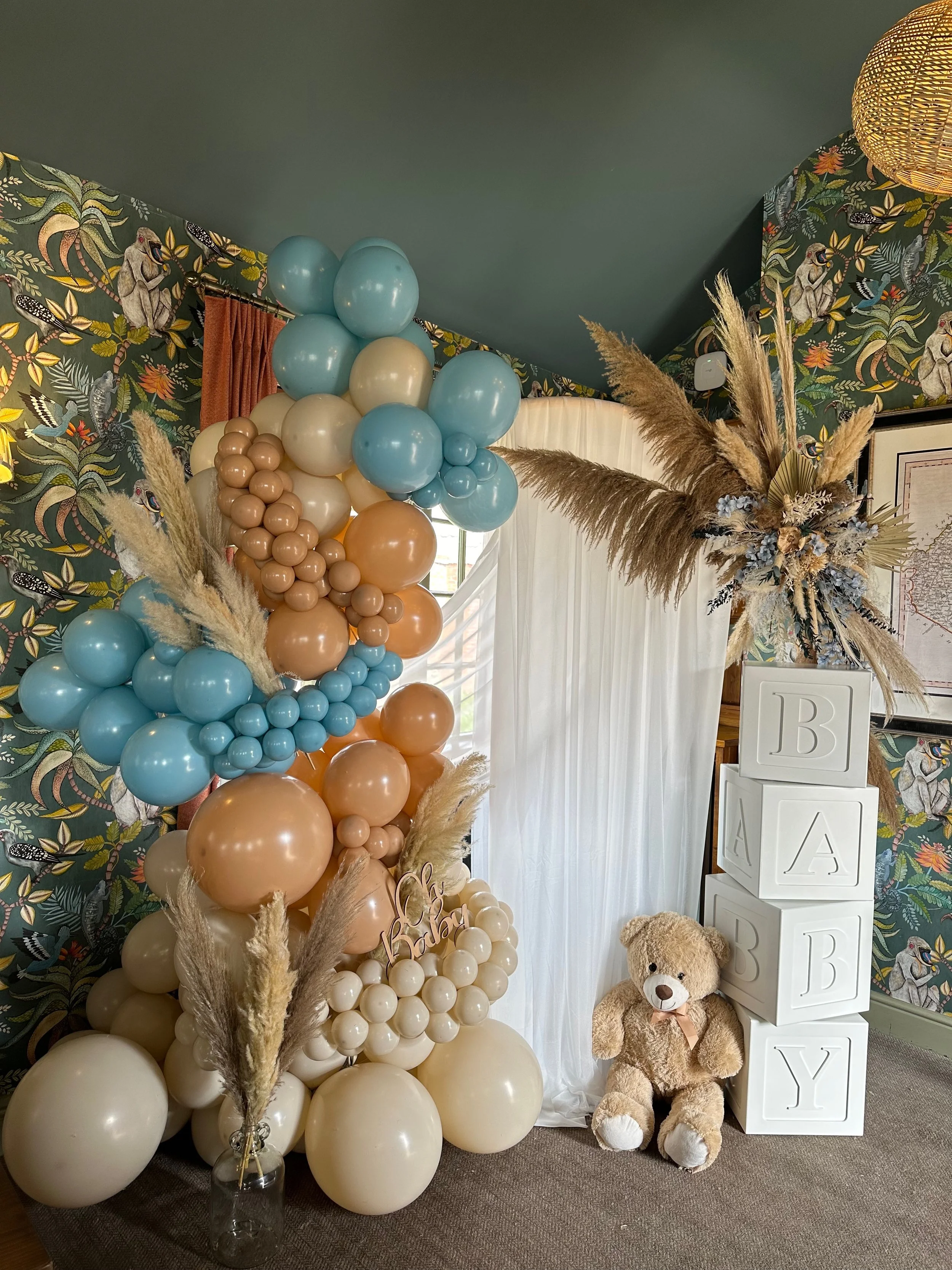 Decorative arrangement for a baby shower, featuring balloons in shades of beige, blue, and peach, pampas grass, a plush teddy bear, and blocks spelling 'BABY' with a white curtain backdrop.