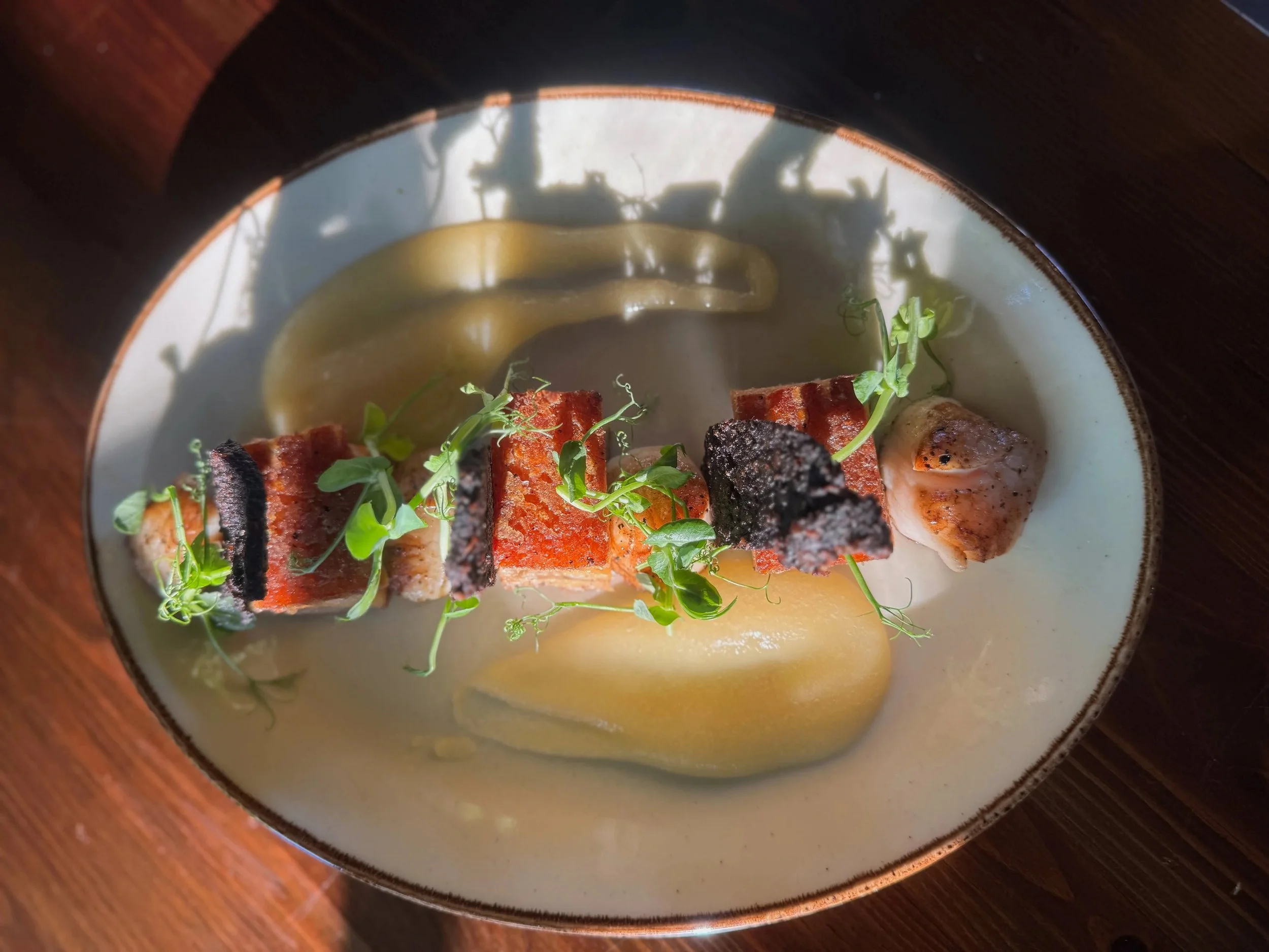 A plated dish featuring grilled salmon topped with microgreens, accompanied by roasted garlic, grilled lemon, and a smear of green sauce, served on a ceramic plate.