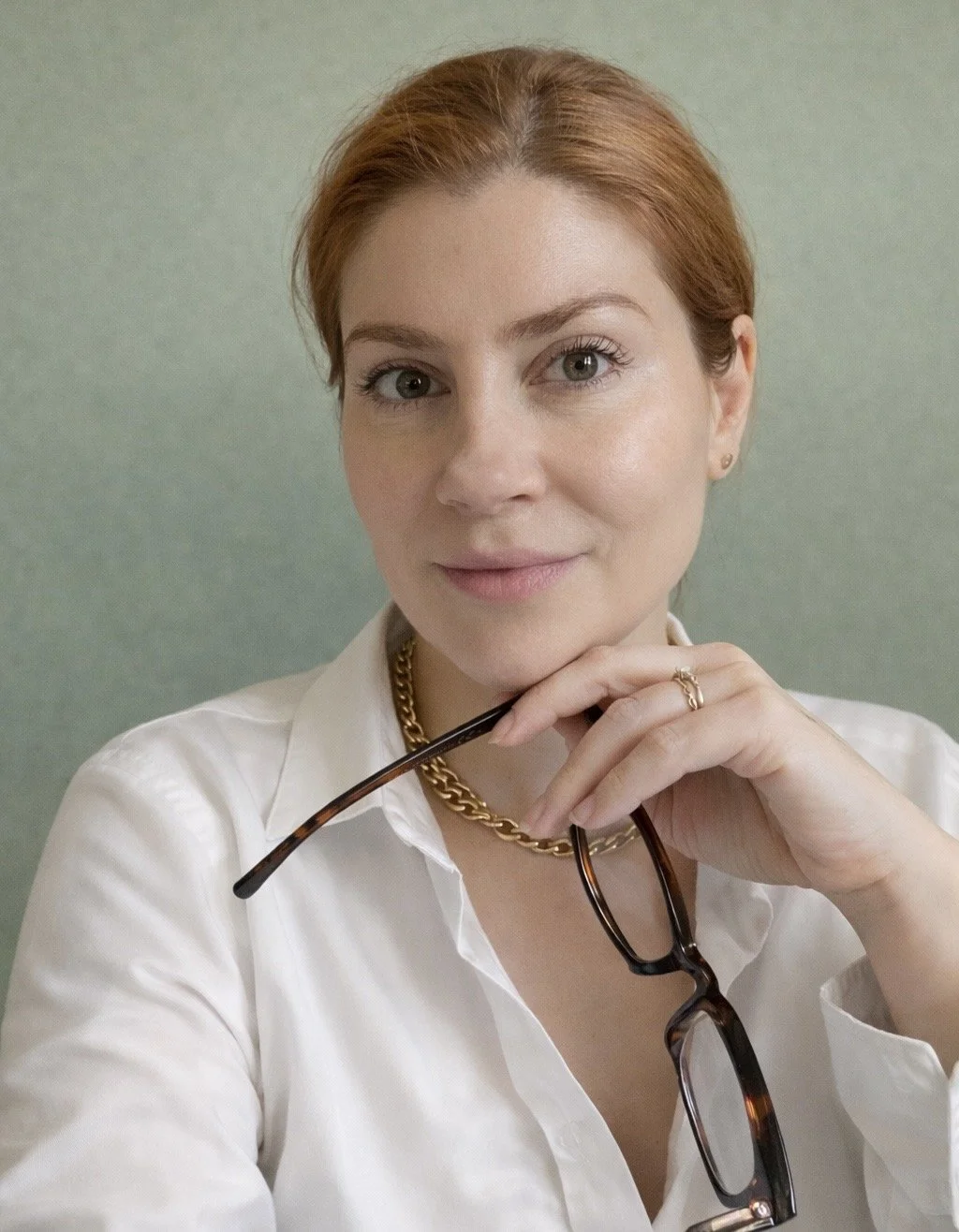 A woman with red hair, wearing a white shirt, gold chain necklace, and holding tortoiseshell glasses, looking at the camera against a green background.