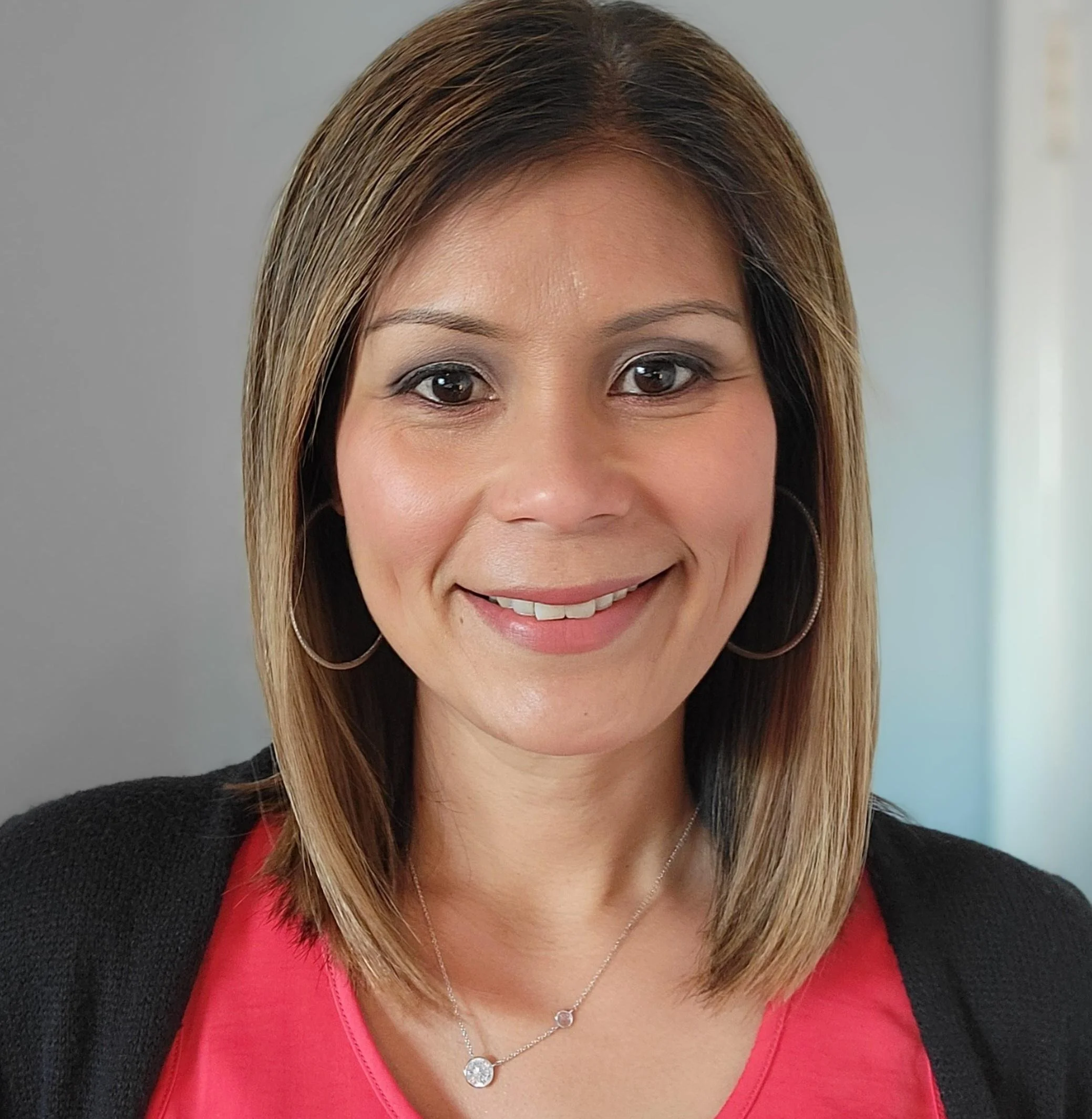 A smiling woman with shoulder-length brown hair and hoop earrings, wearing a pink top and black cardigan, indoors.