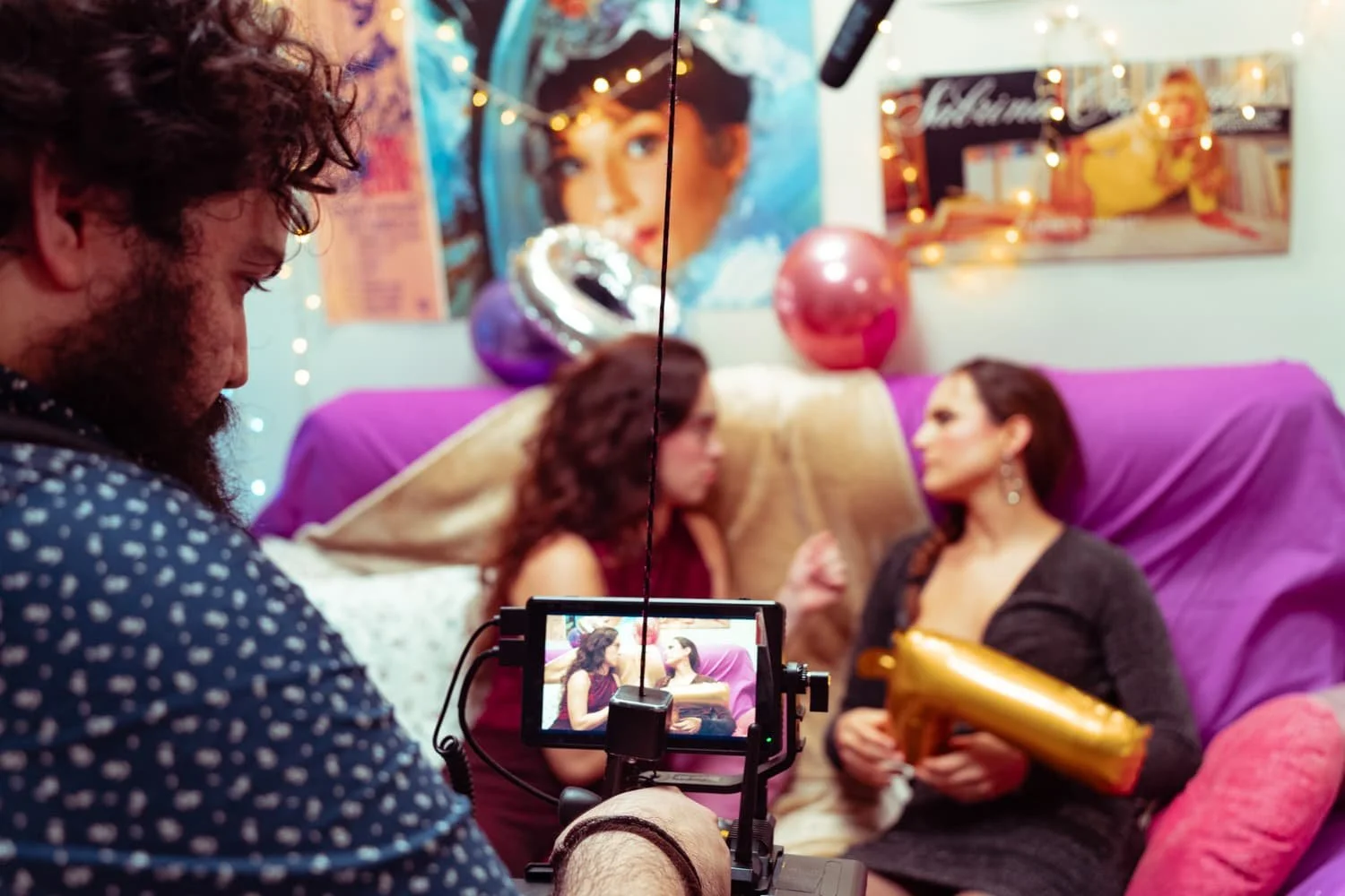 Un hombre filma una escena con dos mujeres en una sala decorada con globos y posters vintage, con luces de tema de fiesta.