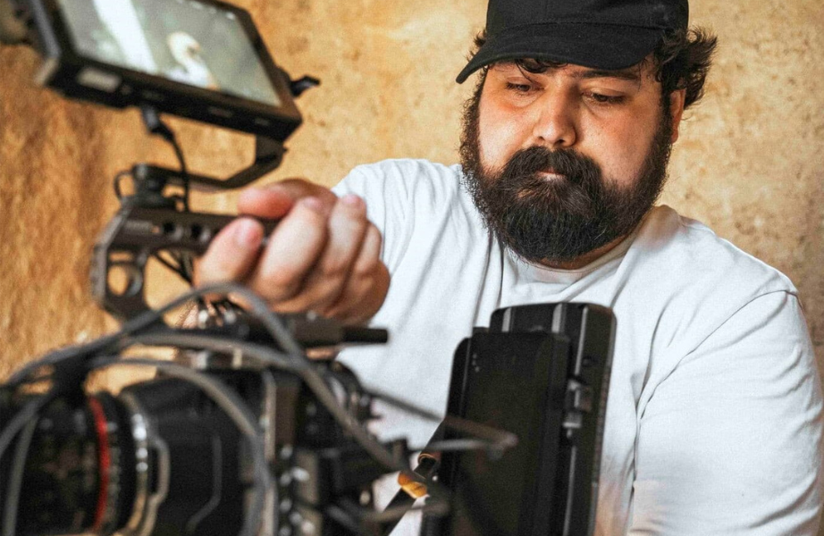 Hombre con barba y gorra ajustando equipo de grabación con cámara en fondo de pared de color arena.