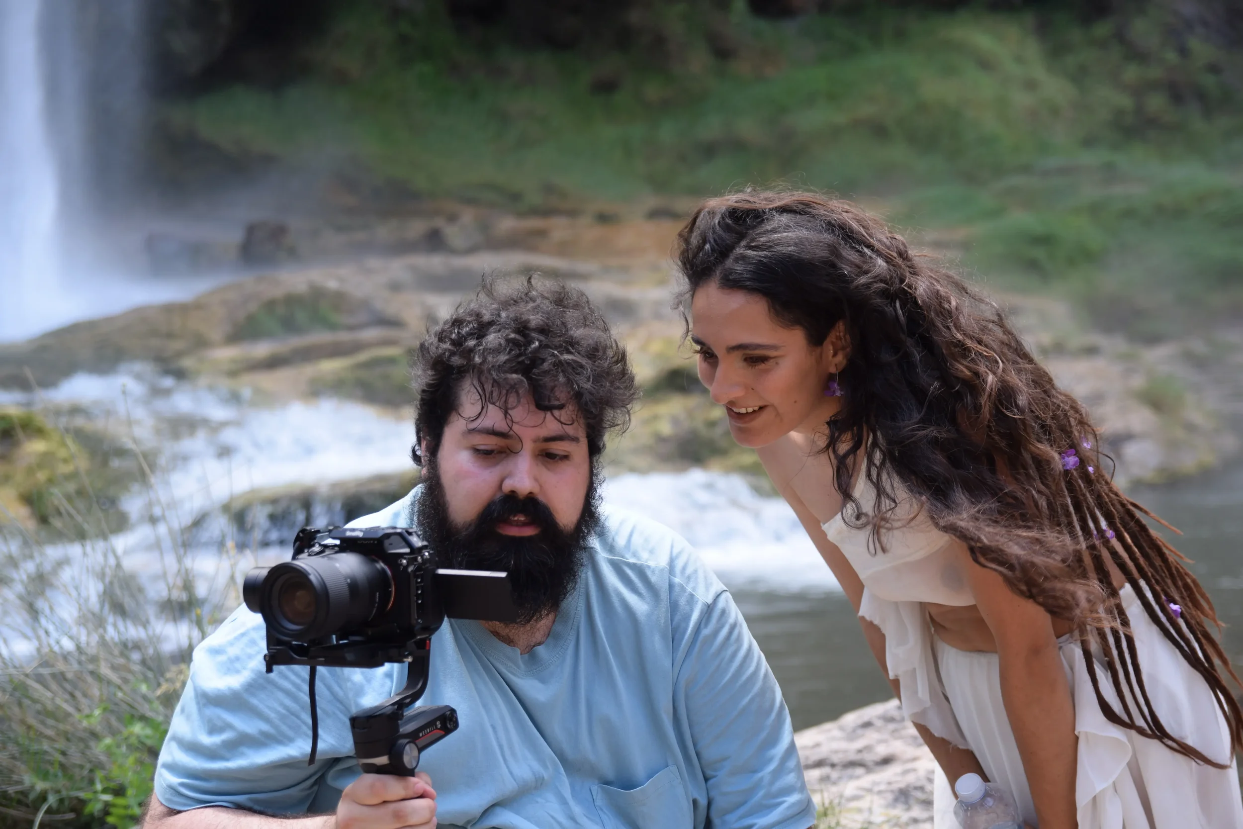 Dos personas, un hombre con barba y una mujer con cabello rizado, observan una cámara de video durante una grabación junto a una cascada en un entorno natural.
