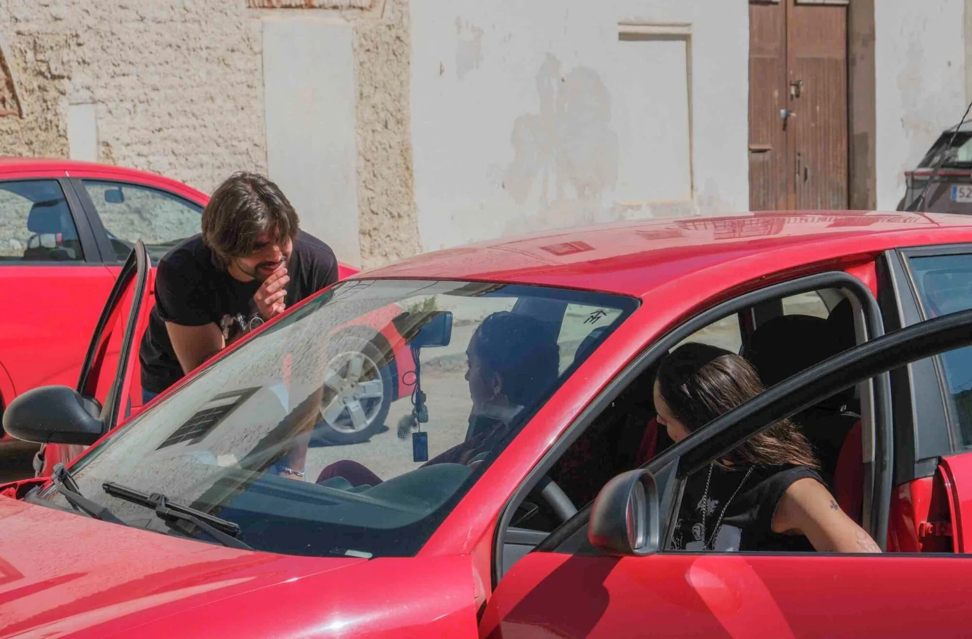 Tres personas interactuando con dos autos rojos en una calle, una mujer en el asiento del conductor y un hombre de pie, ambos riendo.