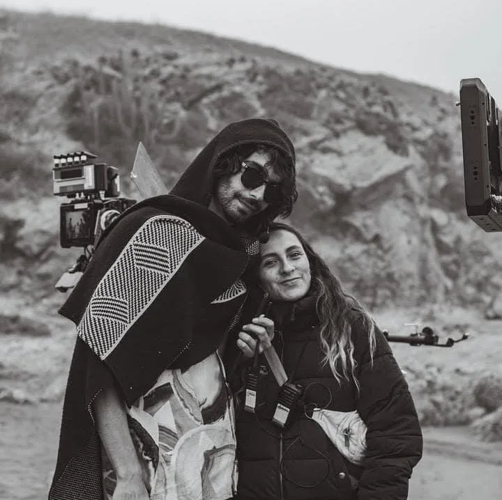 Dos personas, un hombre con gafas oscuras y una chaqueta con capucha, y una mujer con cabello rizado, posando juntos en un paisaje montañoso, en un escenario de filmación.