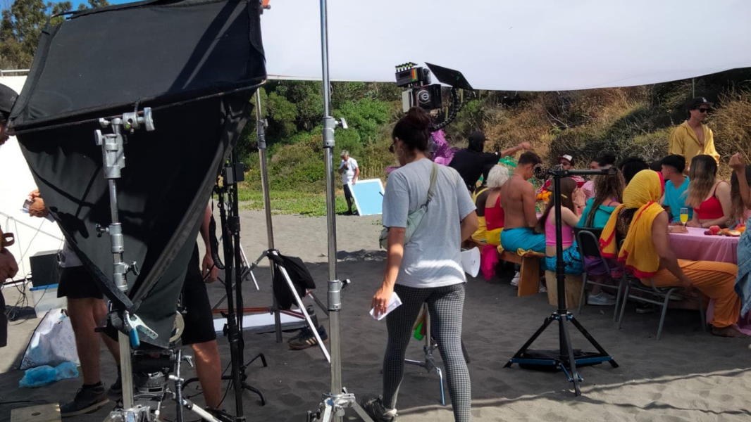 Equipo de filmación grabando una fiesta con muchas personas sentadas en mesas con ropa colorida bajo una lona en la playa.