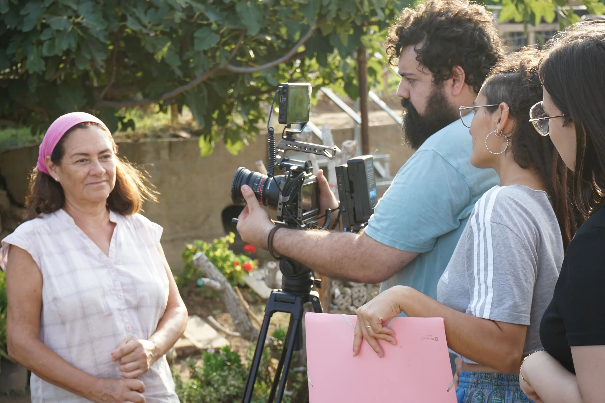 Mujer mayor siendo filmada por un equipo de producción de video en un jardín, mientras otros miembros del equipo sostienen una libreta rosa y una tablet.