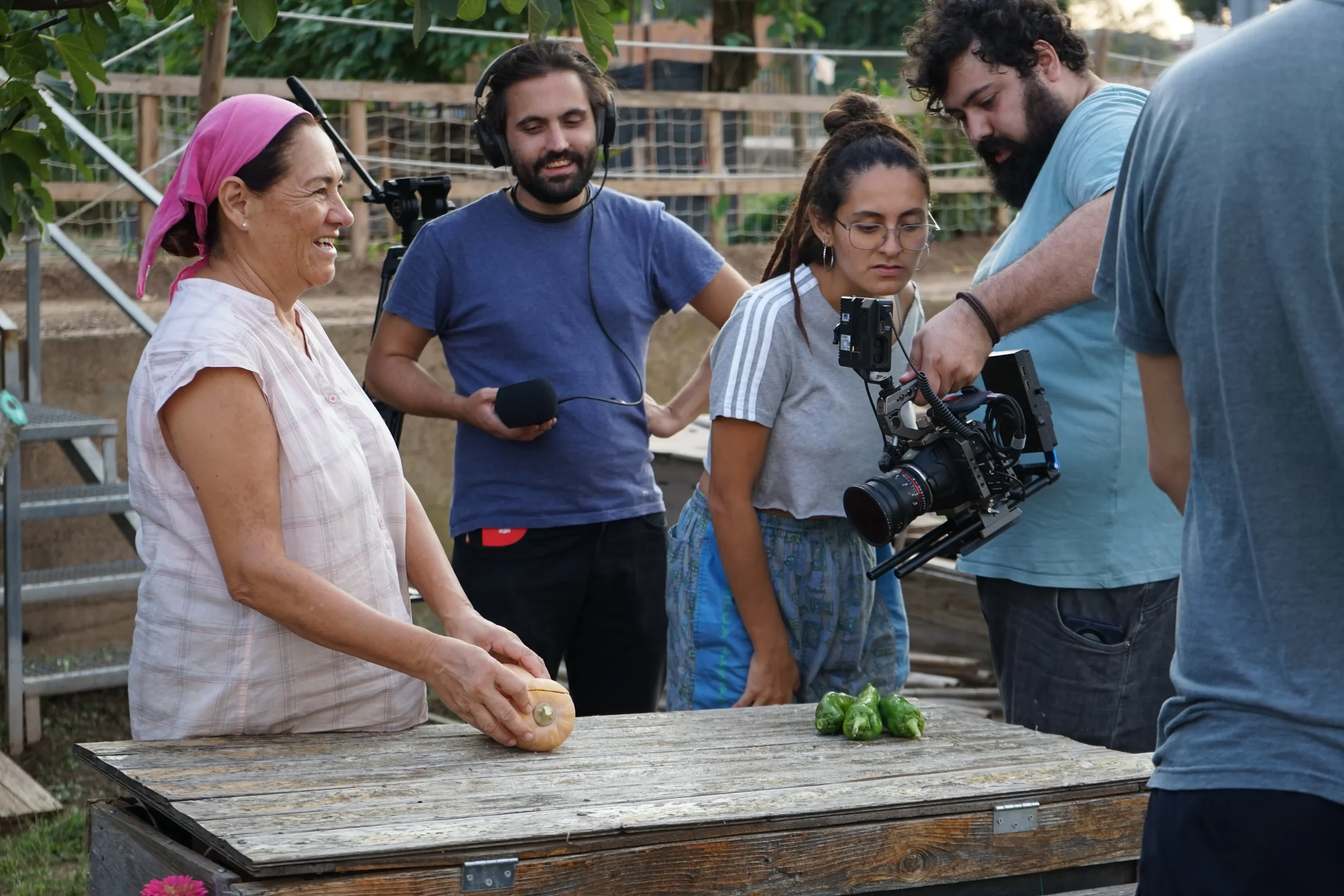 Un grupo de cinco personas trabaja en un huerto, con una mujer mayor que sostiene una calabaza y varias habas frente a ella, mientras dos hombres y una joven observan y uno de los hombres ajusta una cámara de video. Todos parecen estar en un proyecto