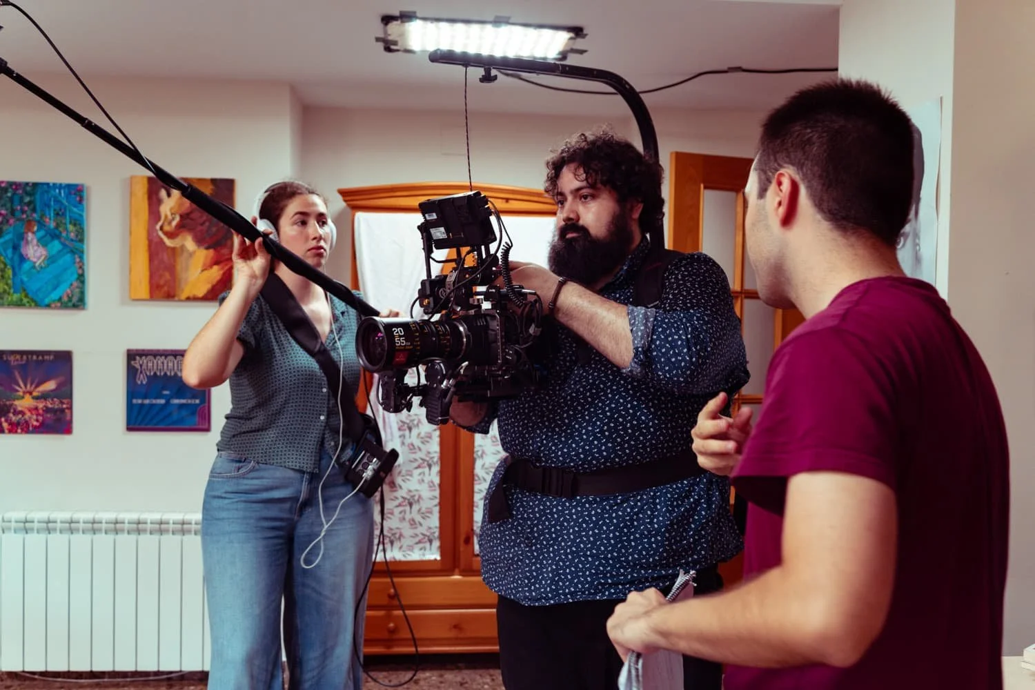 Camarógrafo ajustando una cámara en un set de filmación mientras dos personas lo observan, en una habitación decorada con cuadros y muebles de madera.