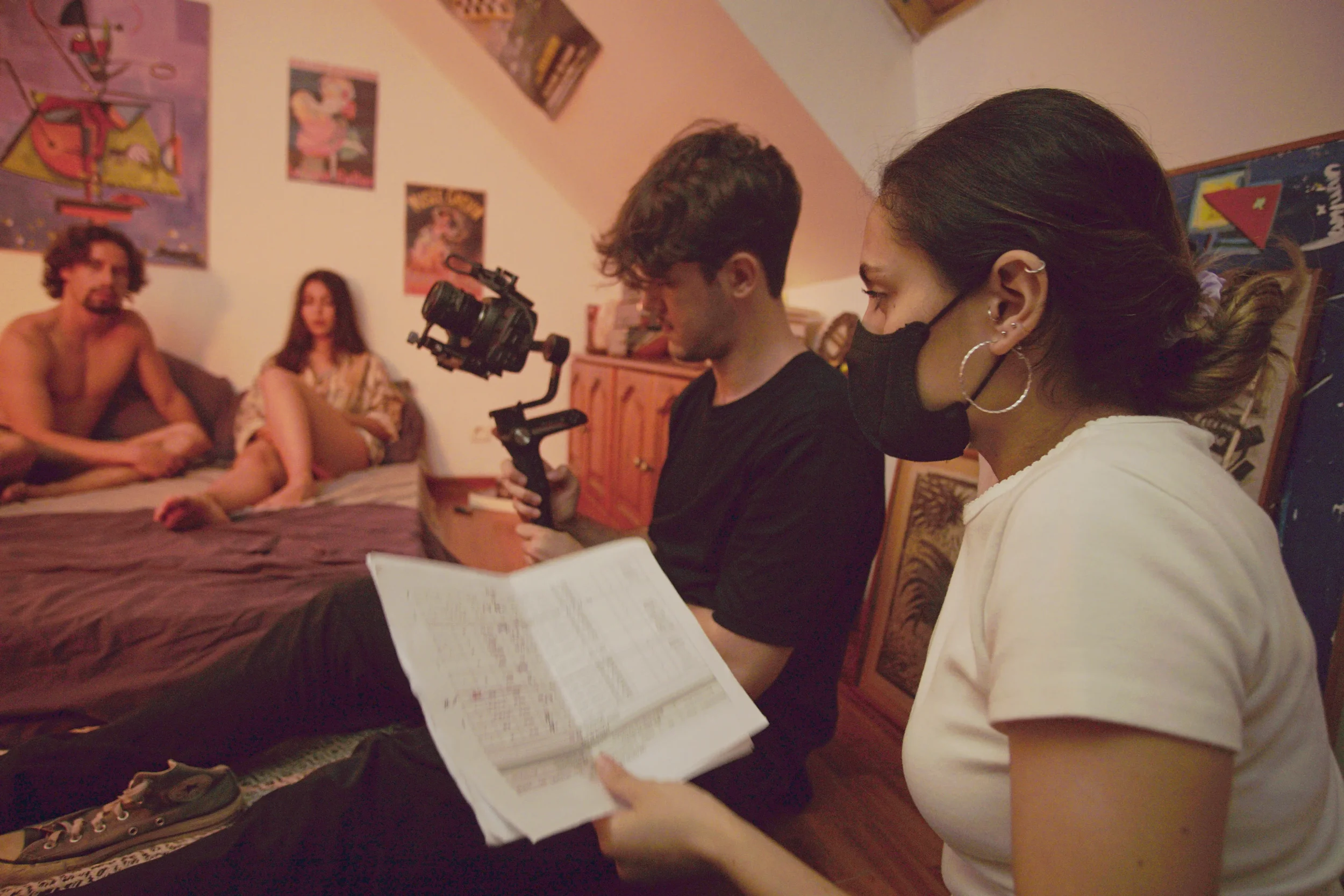 Dos personas en un rodaje, una con máscara y sosteniendo un guion, enfocadas en dos actores en un escenario que parecen estar actuando en un ambiente doméstico