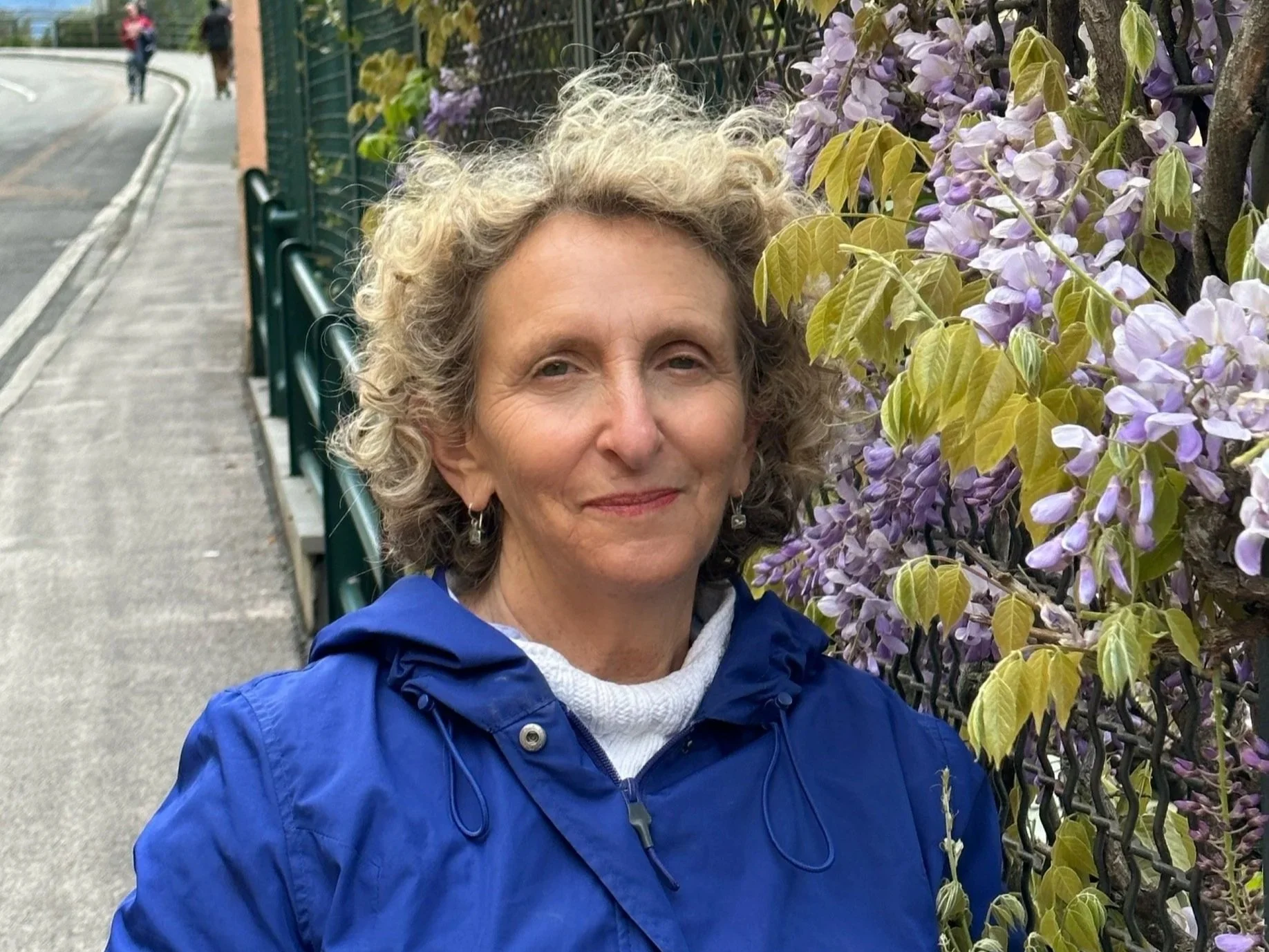 A woman with curly gray hair standing next to a wall of purple wisteria flowers, wearing a blue jacket and earrings, on a sidewalk with a street in the background.