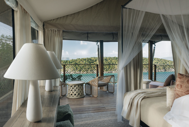 View from inside a tent-style room overlooking a lake, with a balcony, wicker chairs, a side table, a bed with pillows, and curtains.
