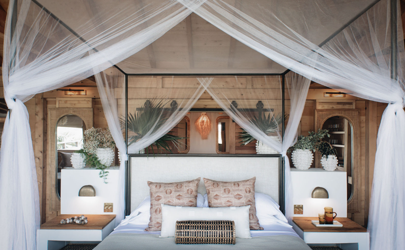 Bedroom with four-poster canopy bed, white mosquito netting, beige pillows, wooden nightstands with lamps, and decorative plants in white vases.