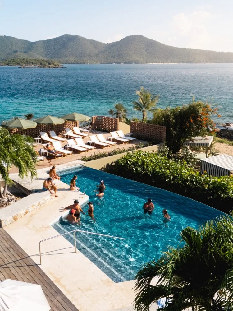 People relaxing and swimming in a pool by the ocean with mountains in the background, surrounded by palm trees and lounge chairs.