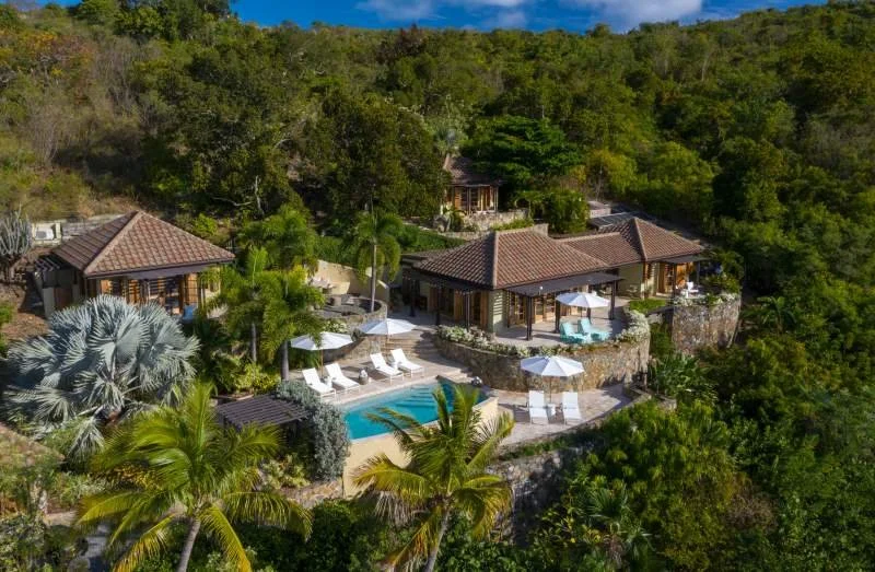 A luxurious tropical estate featuring multiple villas with red-tiled roofs, a swimming pool with lounge chairs and umbrellas, surrounded by lush greenery and palm trees.
