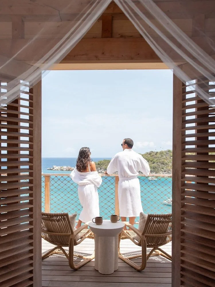 A couple in white robes standing on a balcony, facing the ocean, with a small table and two chairs in the foreground.