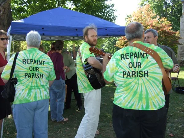people singing with guitars wearing tshirts that say "repaint our parish" as a response to hecklers wearing tshirts that say "repent or perish"