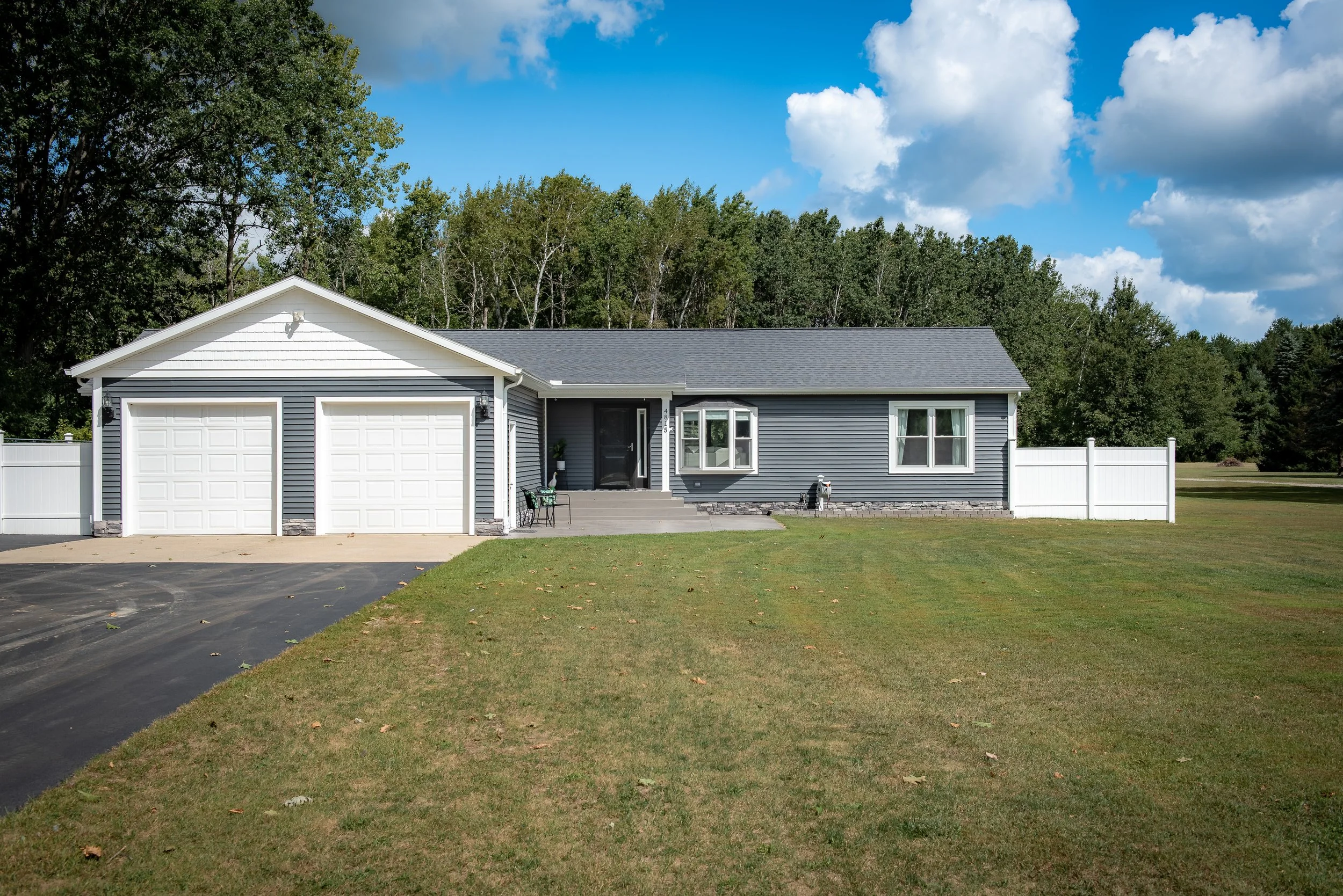 Front view of a suburban house with a two-car garage, a well-maintained lawn, and a white fence on both sides, under a partly cloudy sky.