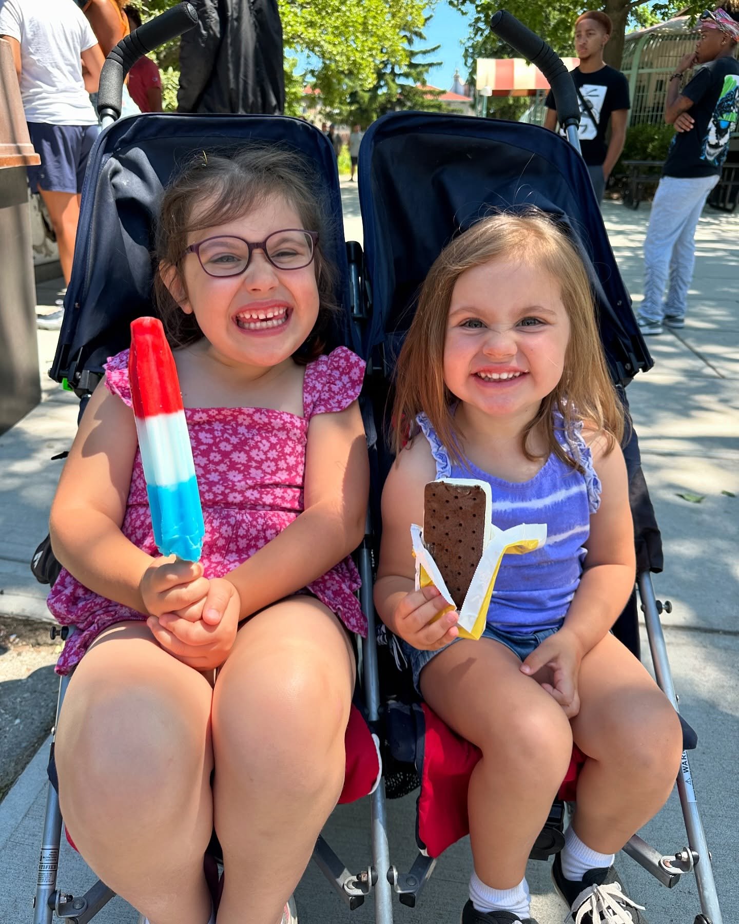 Two young girls sitting in stroller seats outdoors, smiling and holding treats. The girl on the left has glasses and is holding a red, white, and blue popsicle, while the girl on the right is holding a large Oreo cookie. Other children are visible in the background.