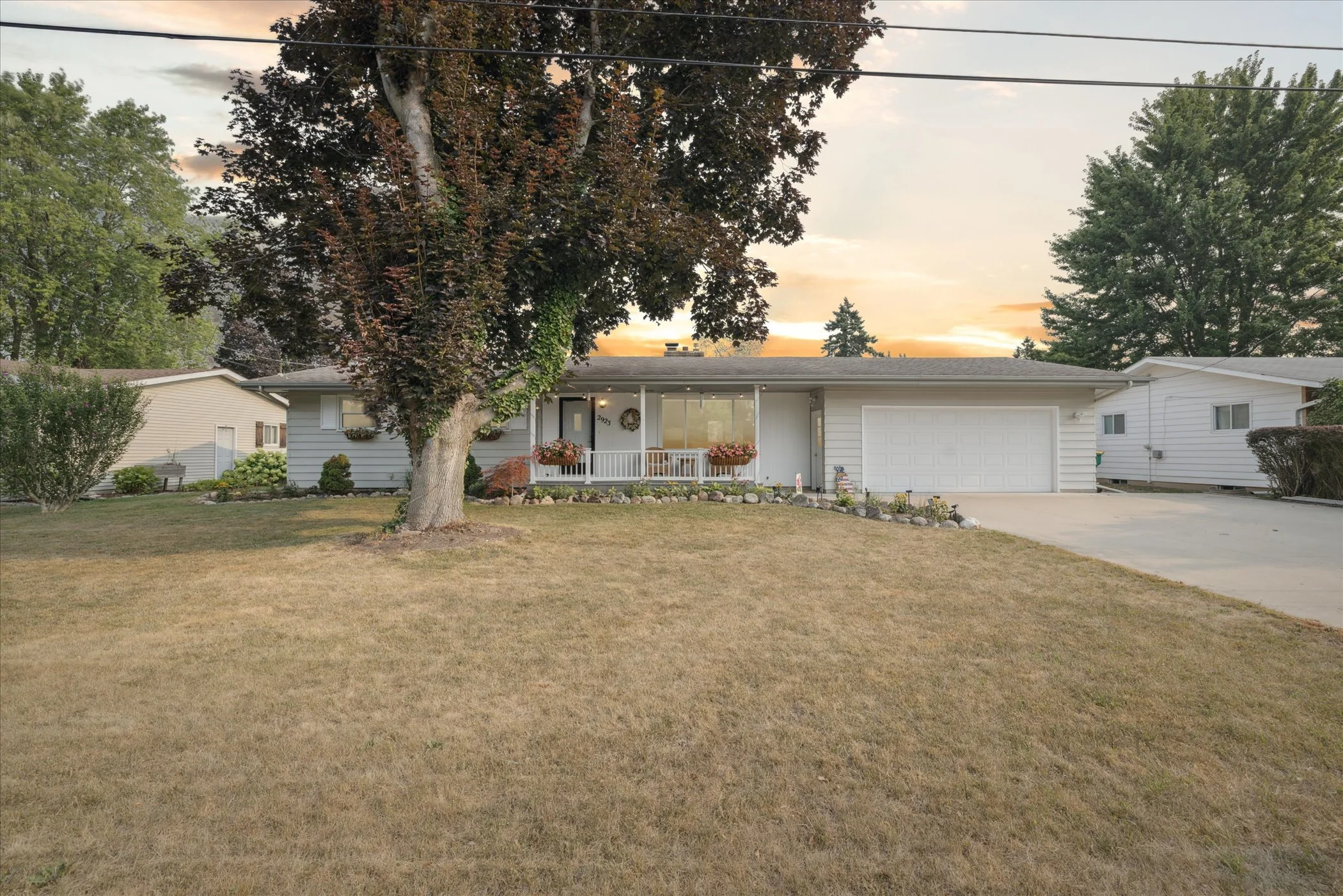 A single-story house with white siding, a two-car garage, front porch with hanging flower baskets, and a large front yard with a tree and some landscaping at sunset.