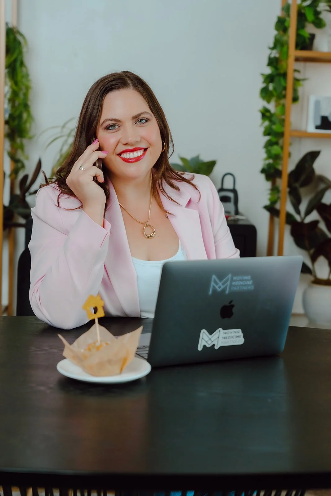 A woman with brown hair, smiling, wearing a pink blazer, sitting at a black table with a laptop and a small plate of food.
