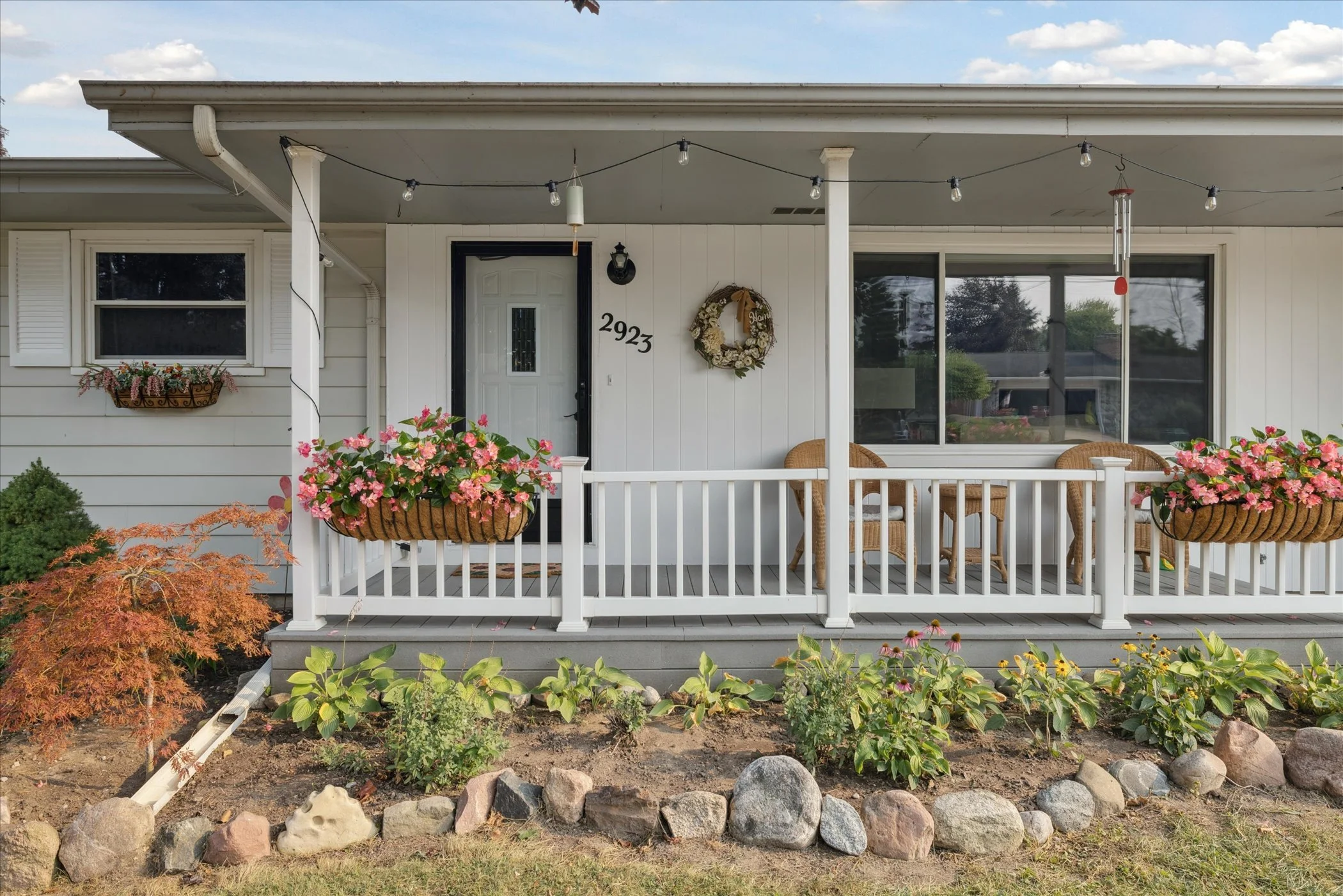 Front porch of a house with white railing, flower baskets, and outdoor seating, decorated with a wreath and wind chimes, and a landscaped front yard with rocks and plants.