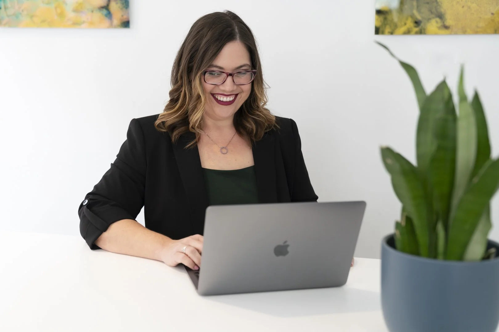 A woman with shoulder-length brown hair, glasses, and a dark blazer is sitting at a white desk, smiling while working on a silver MacBook laptop. There is a large green plant in a blue pot in front of her on the desk, and colorful artwork hanging on the white wall behind her.