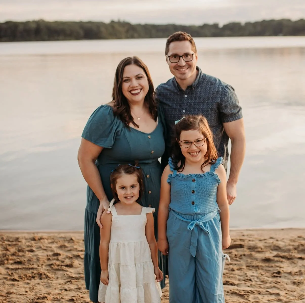 A family of four, including two young girls and two adults, standing on a sandy beach near a body of water with trees in the background during sunset, all smiling at the camera.