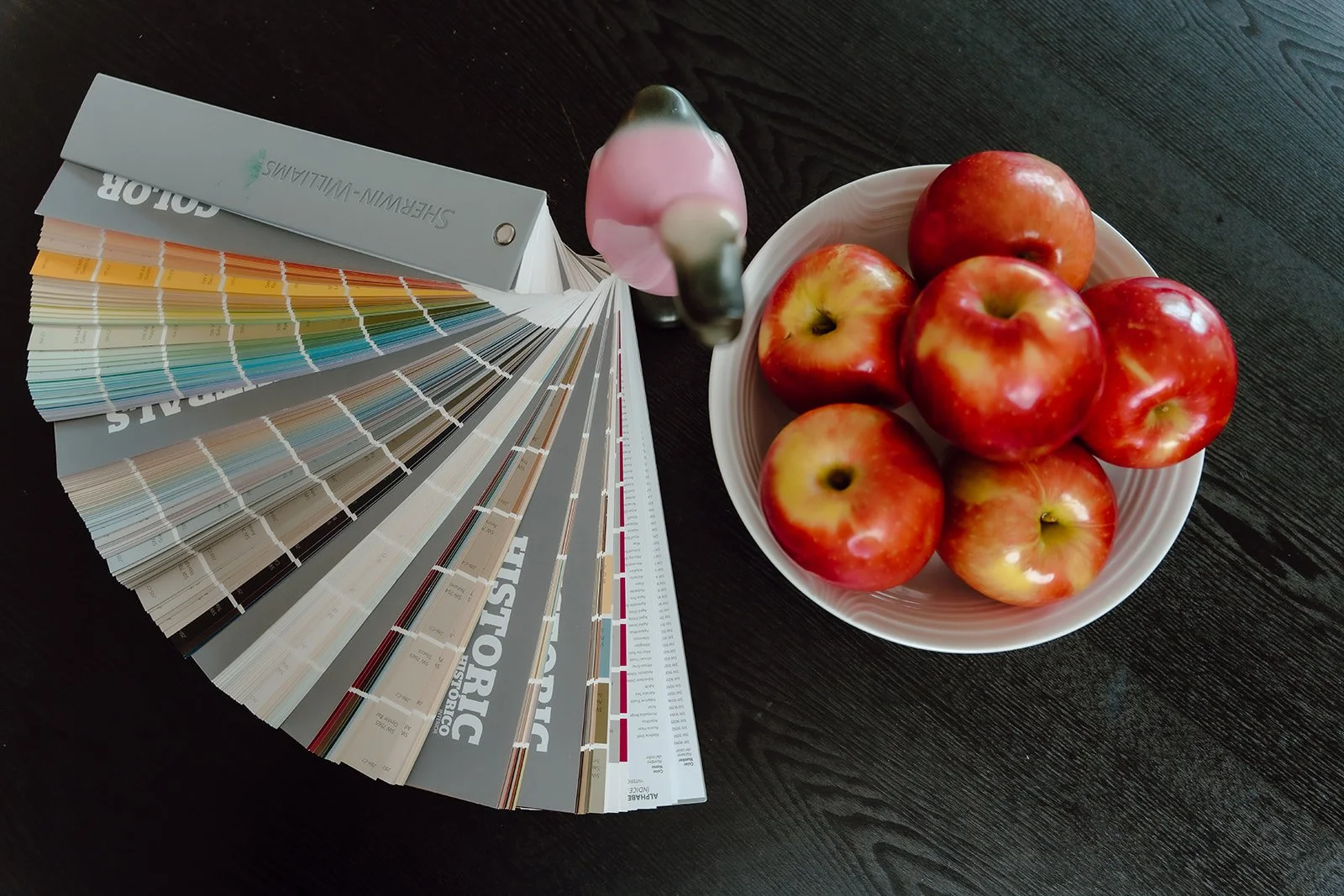 Color swatches fan deck, a pink and gray paint sample, a white bowl of red apples, on a dark wood table.