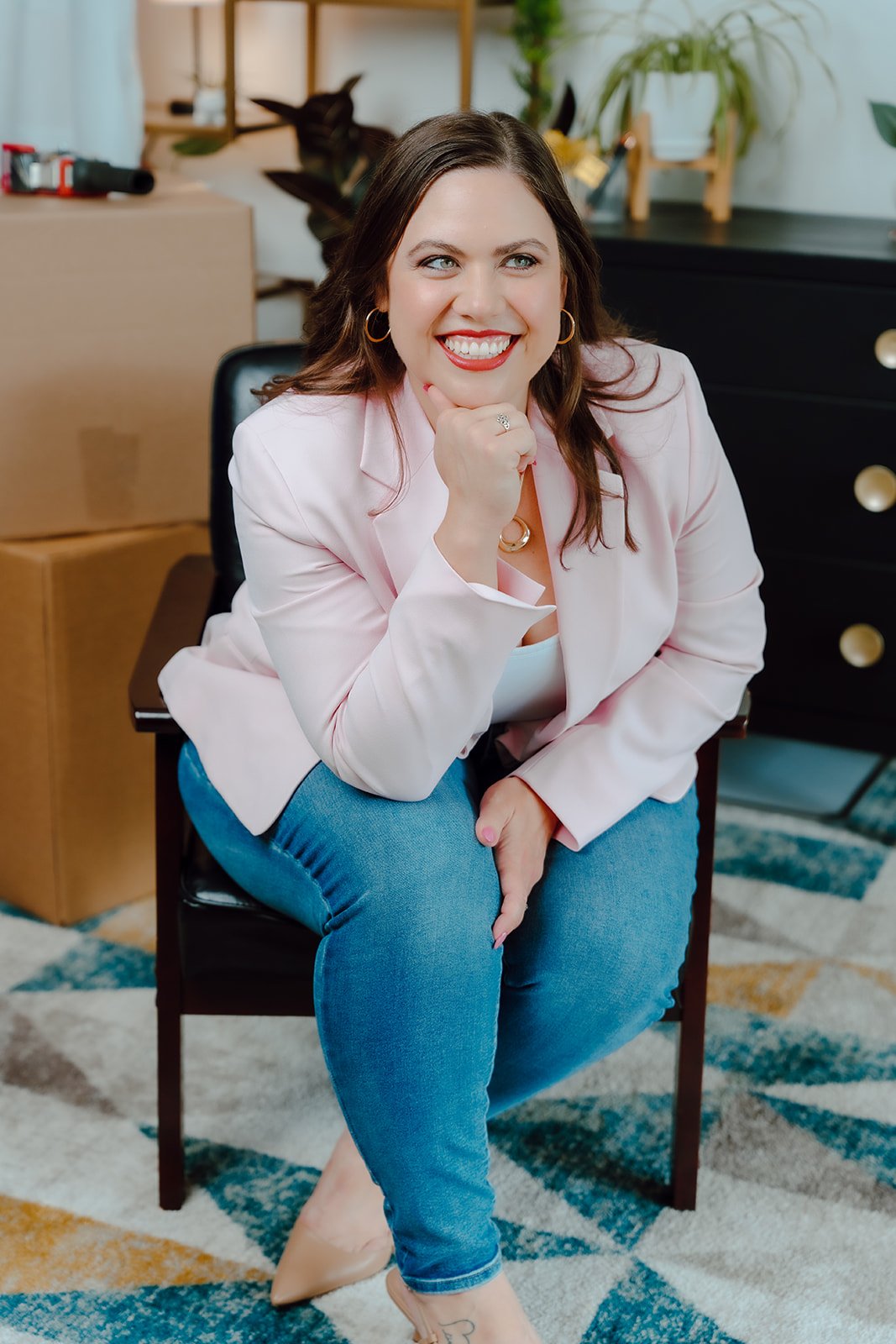 A woman with brown hair, wearing a pink blazer, white top, and blue jeans, smiling and sitting on a black chair in a room with plants and boxes.