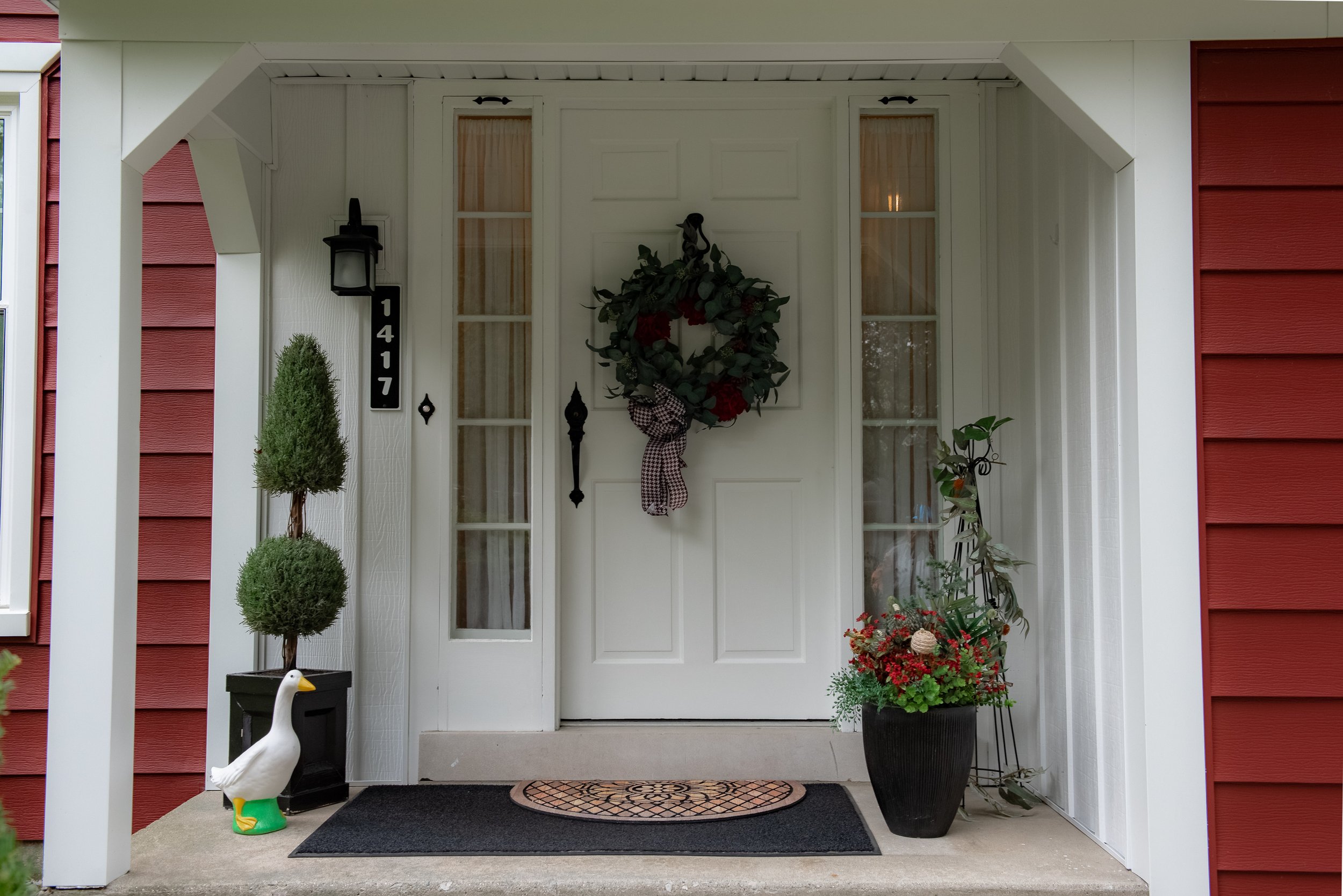 Front porch of a house with a white door decorated with a wreath, flanked by two tall windows, and adorned with potted plants and garden decorations.