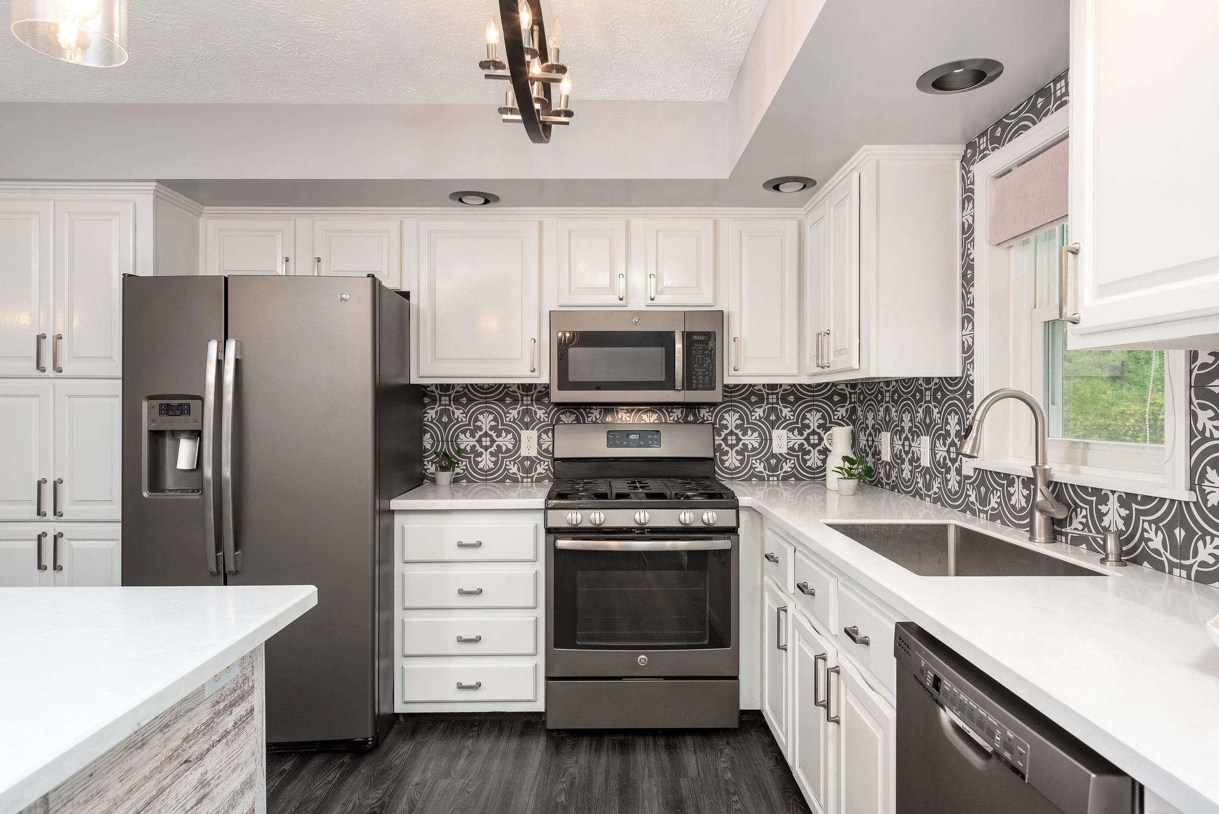 Modern kitchen with white cabinets, gray patterned backsplash, stainless steel refrigerator, oven, microwave, sink under window, and dark wood flooring.