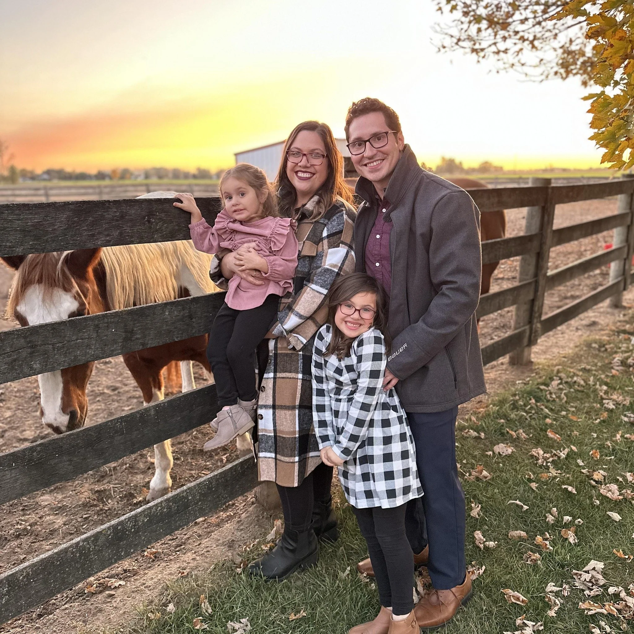 A family of four, including two young girls and their parents, standing close together at a farm during sunset. The family is smiling and posing near a wooden fence with horses in the background.