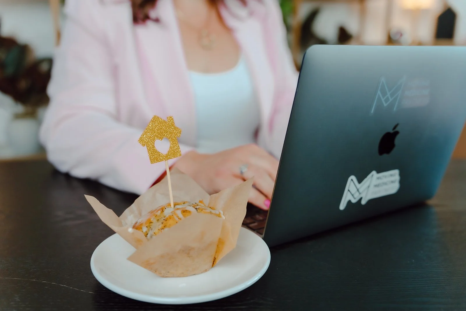 A person working on a laptop at a table, with a cupcake decorated with a gold glitter house and heart pick in the foreground.