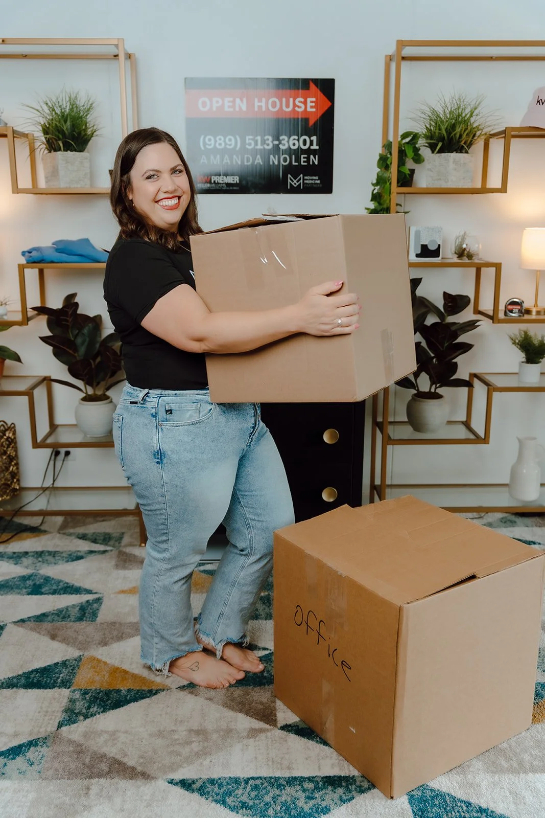A woman with dark brown hair, wearing a black t-shirt and ripped jeans, smiling and holding a large cardboard box inside a room with decorative wall shelves and plants. There is a second box on the floor labeled "OFFICE" and a sign behind her that says "Open House" with a phone number and name.