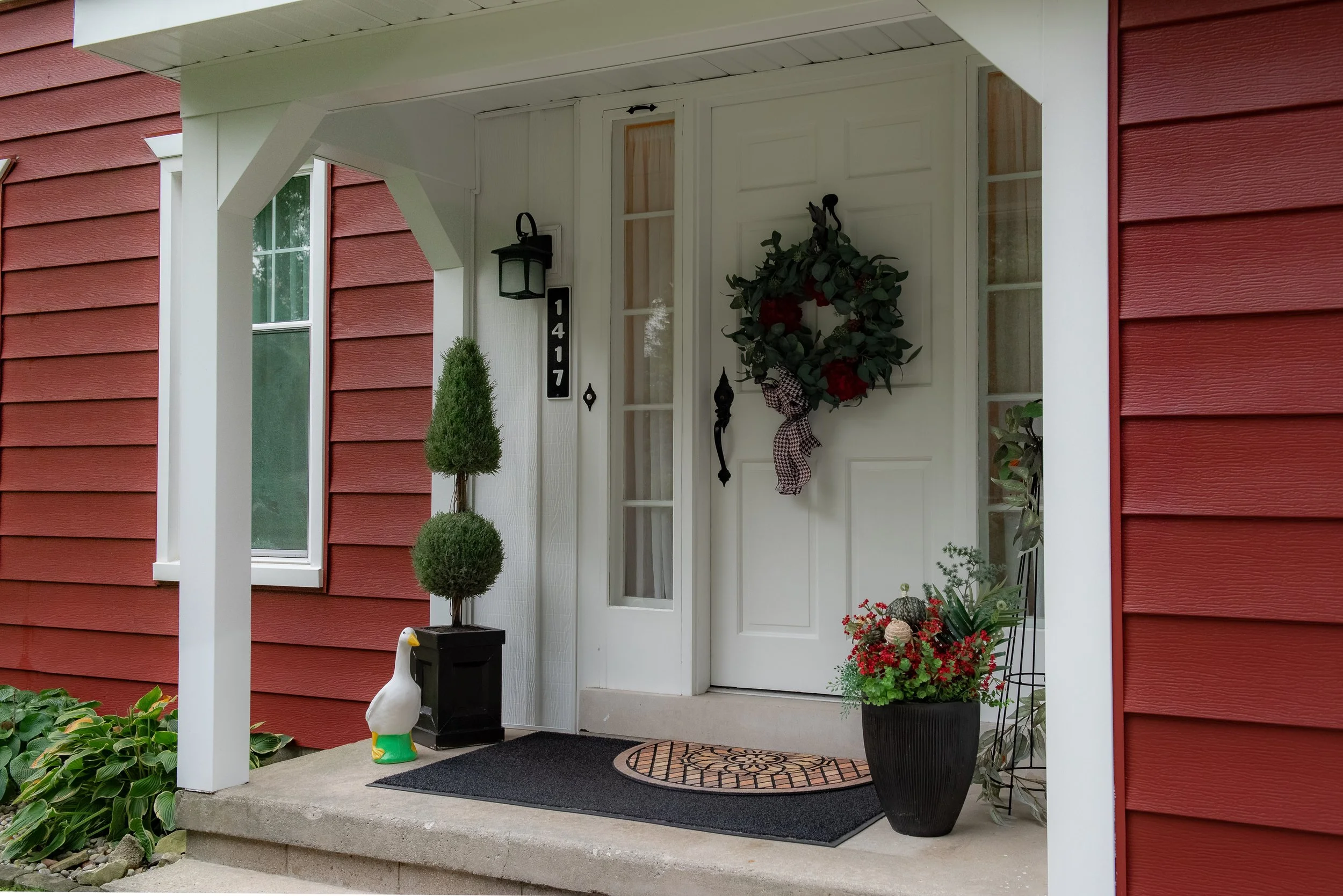 Front porch of a house with red siding, white door with a wreath, potted plants, and a decorative duck statue on the step.