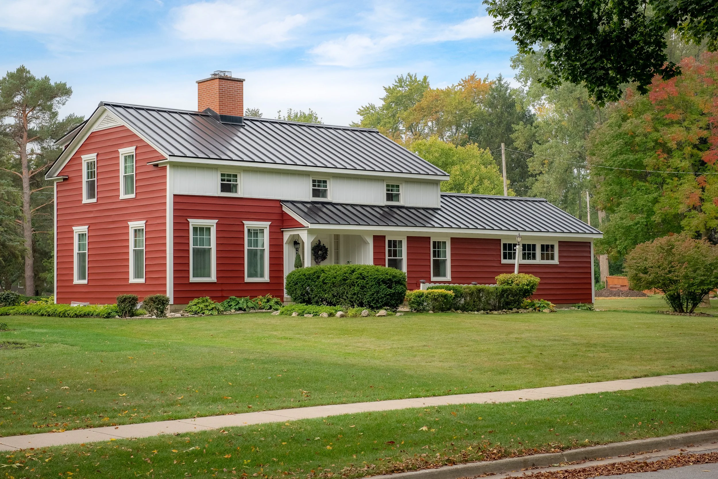 A two-story house painted red with white trim, metal roof, and a front porch, surrounded by a well-maintained lawn and trees with fall foliage.