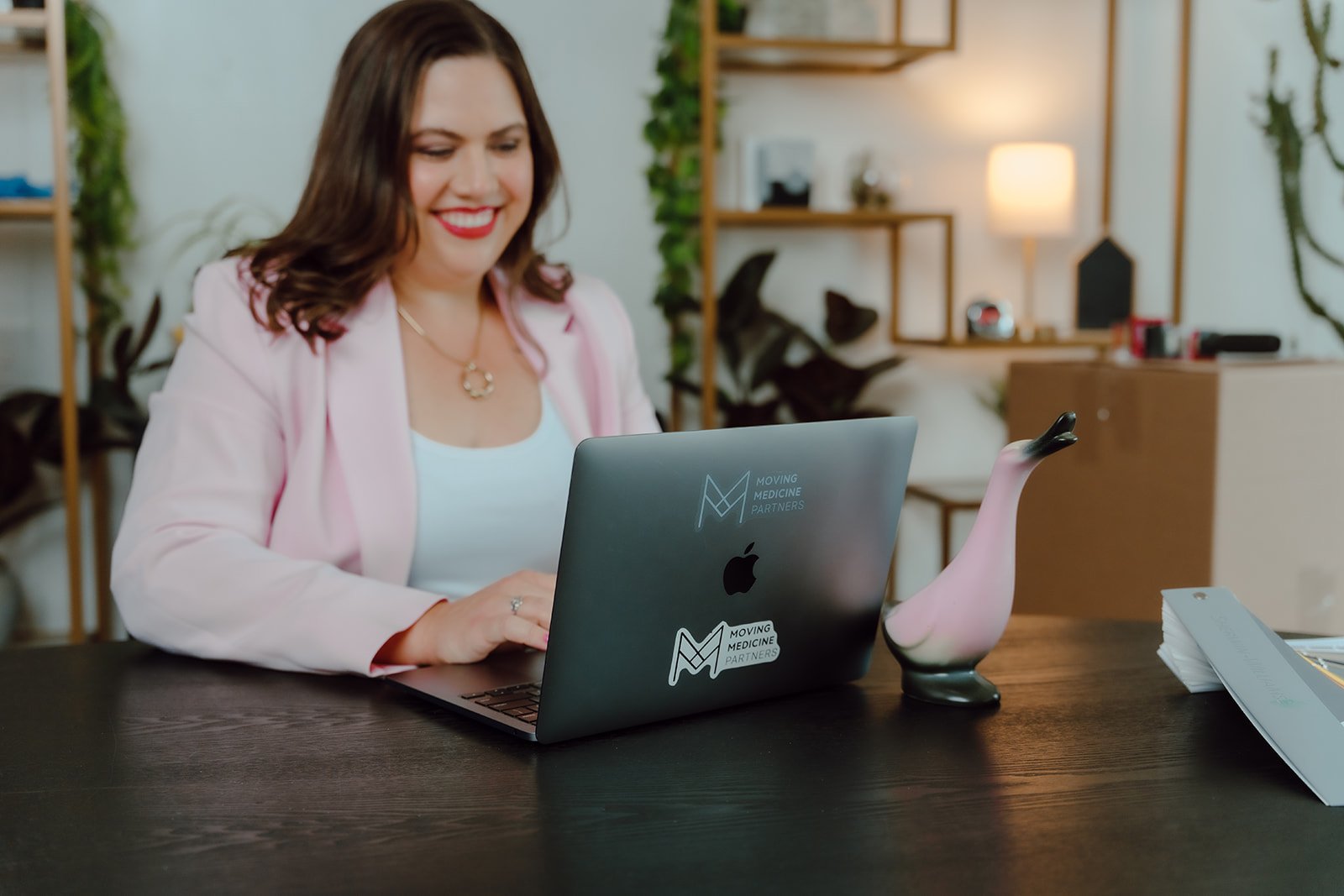 A woman sitting at a desk, smiling while using a laptop with a sticker that reads 'Moving Medicine Partners.' A decorative duck figurine wearing a pink hat and a DVD or book are on the desk. The background features a bookshelf with plants and a lamp, in a modern office or home workspace.